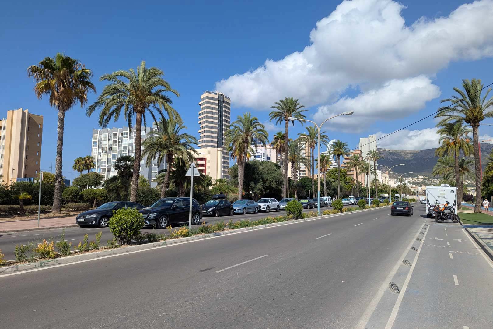 Uitzicht op een hoofdweg in Calpe met palmbomen, een fietspad en hoogbouw, wat de stedelijke omgeving en voorzieningen nabij de woning illustreert.