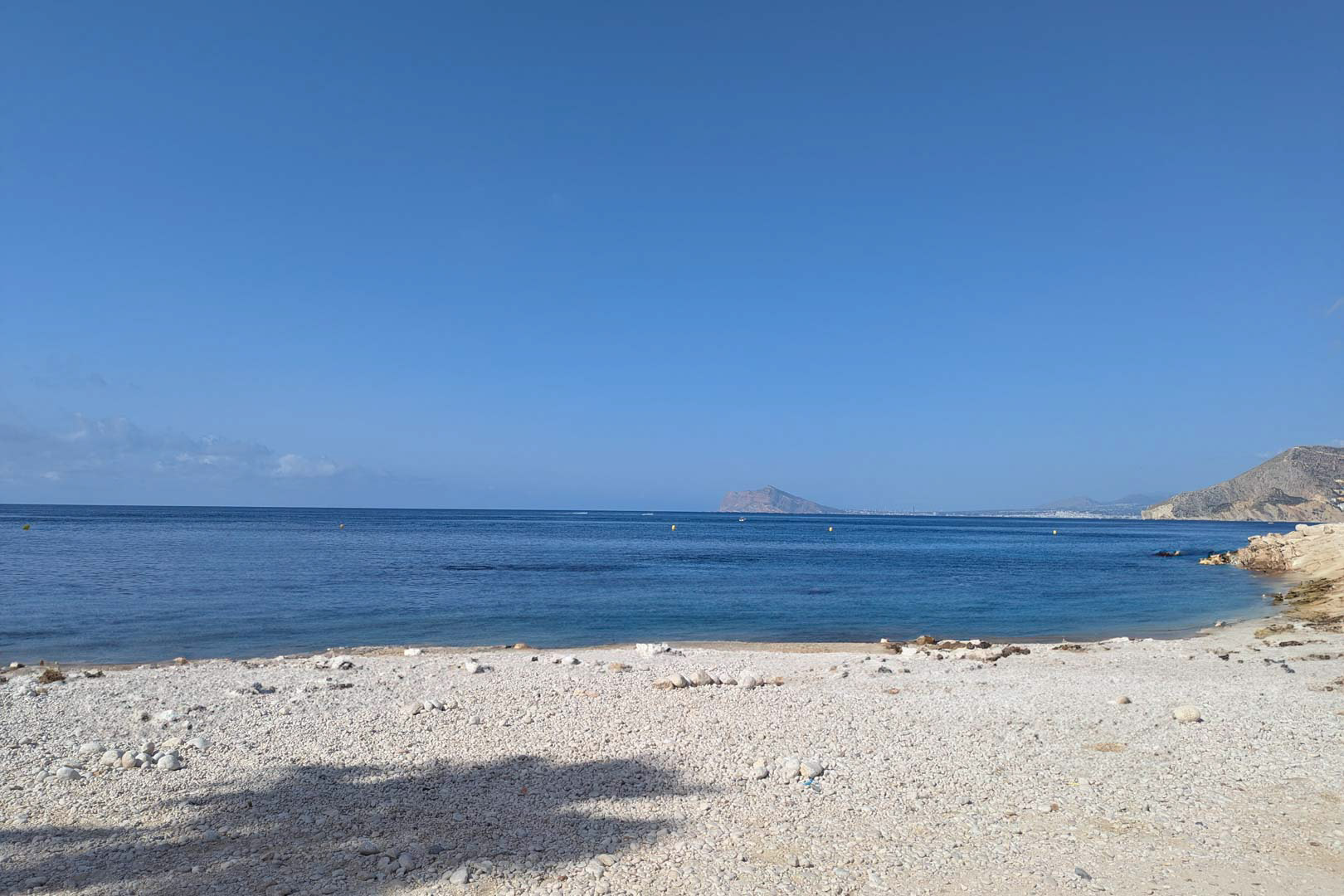 Uitzicht op een witte kiezelbaai met rustig blauw water in Calpe, met de mediterrane kustlijn en verre bergen onder een heldere hemel.
