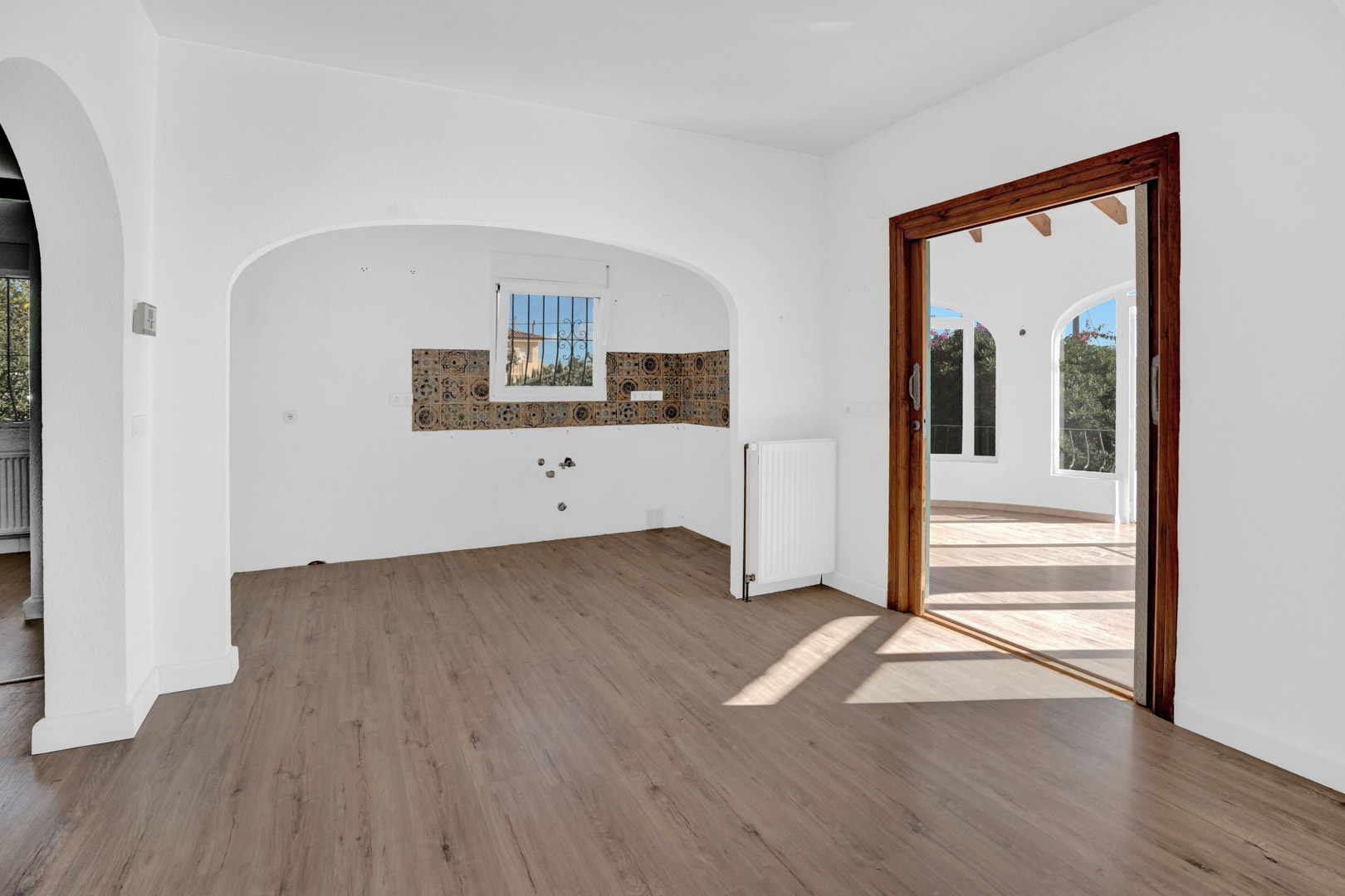 Woonkamer met laminaatvloer, radiator en een keukenhoek met decoratieve tegels. Een houten schuifdeur biedt toegang tot het lichte glazen terras.