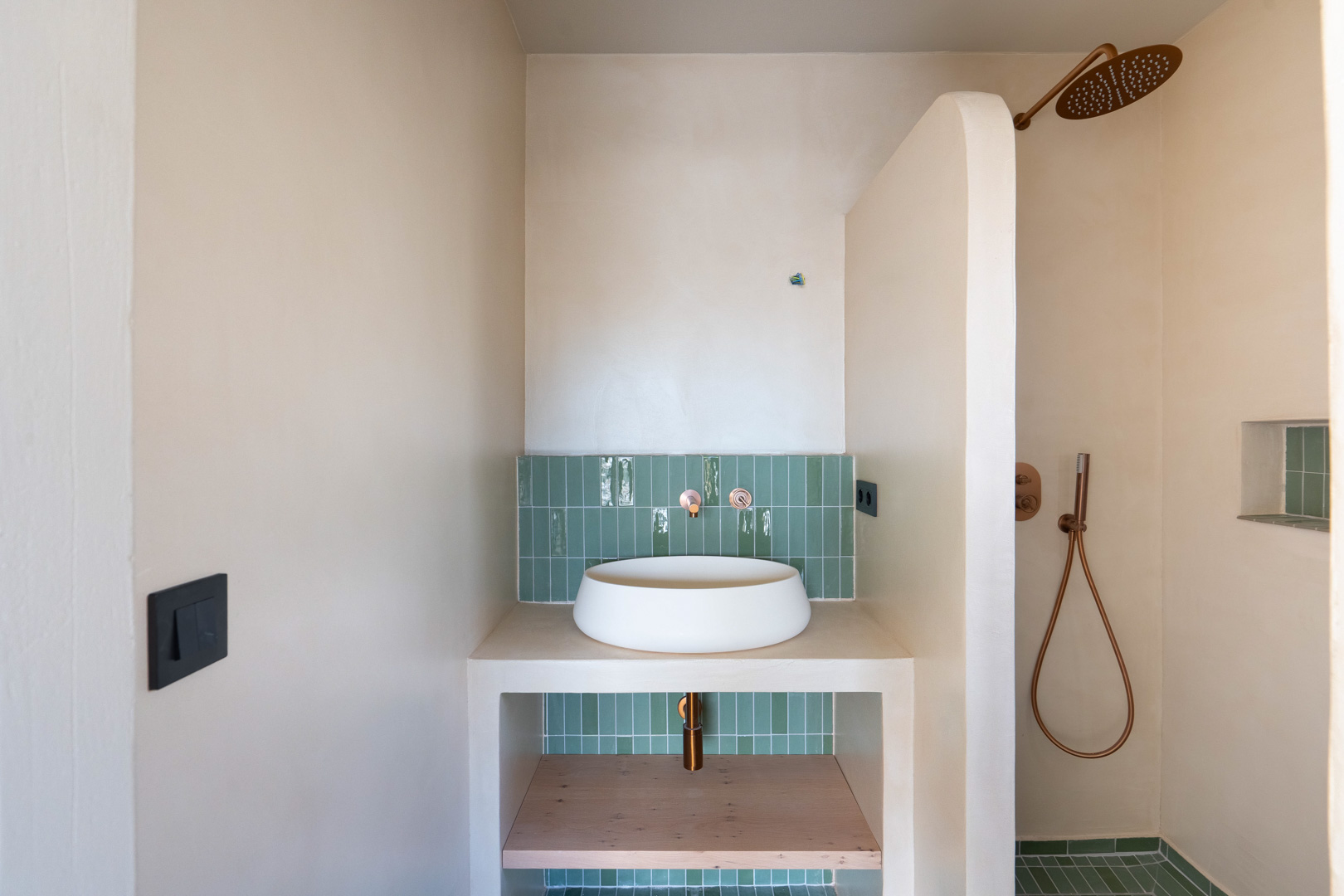 Badkamer met opzetwastafel, koperen kranen en groene verticale tegels. Wanden in leemstuc en een inloopdouche met een kenmerkende gebogen scheidingswand.