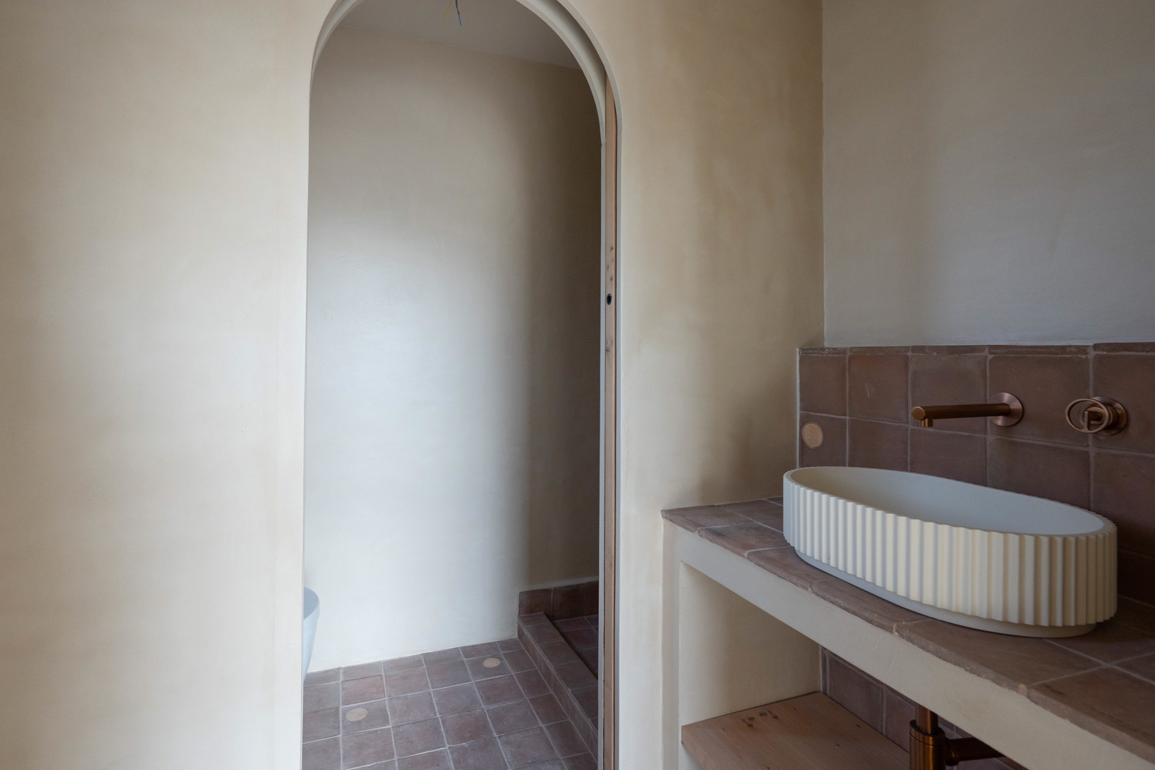 Badkamer met wanden in leemstuc, een geribbelde opzetwastafel en bronzen inbouwkranen. Een stijlvolle boogdoorgang verbindt de ruimte met de terracotta vloer.