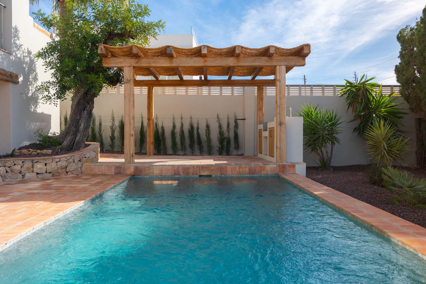 Buitenzwembad met terracotta omlijsting en een rustieke houten pergola met buitenkeuken, omgeven door een mediterrane tuin met olijfbomen en cipressen.