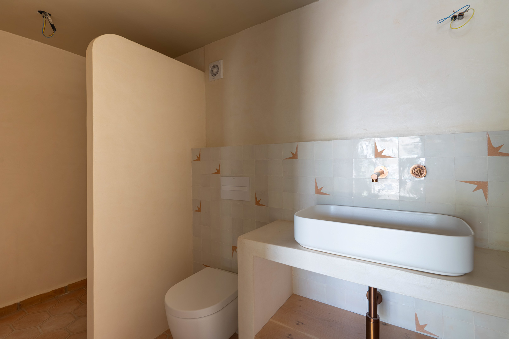 Designbadkamer met opzetwastafel op een gemetseld blad, koperen inbouwkranen en wanden van leemstuc. Voorzien van een hangtoilet en authentieke terracotta vloer.