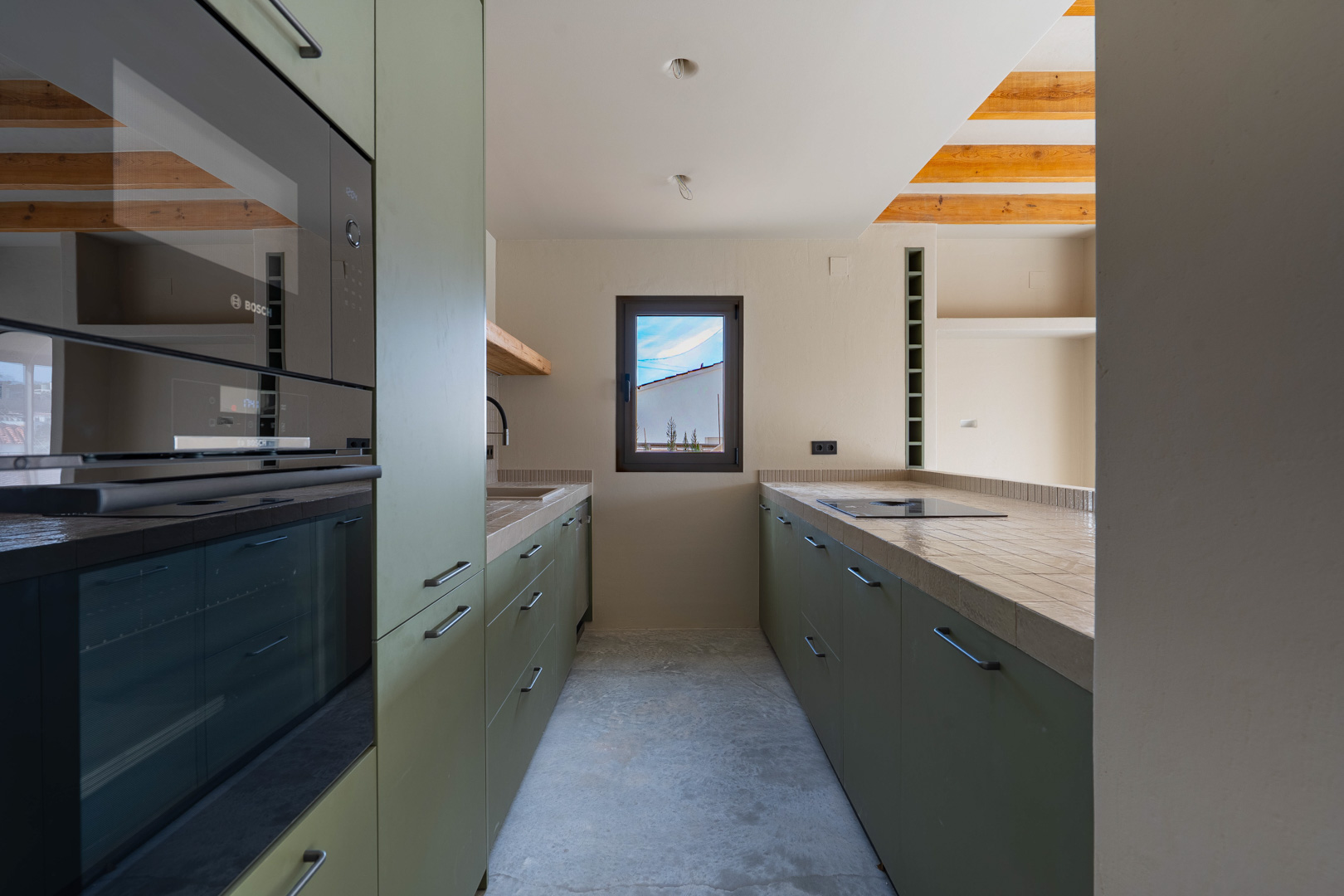Designkeuken met olijfgroene kasten, geïntegreerde Bosch-apparatuur en betegelde werkbladen. Voorzien van houten plafondbalken en een gepolijste betonvloer.