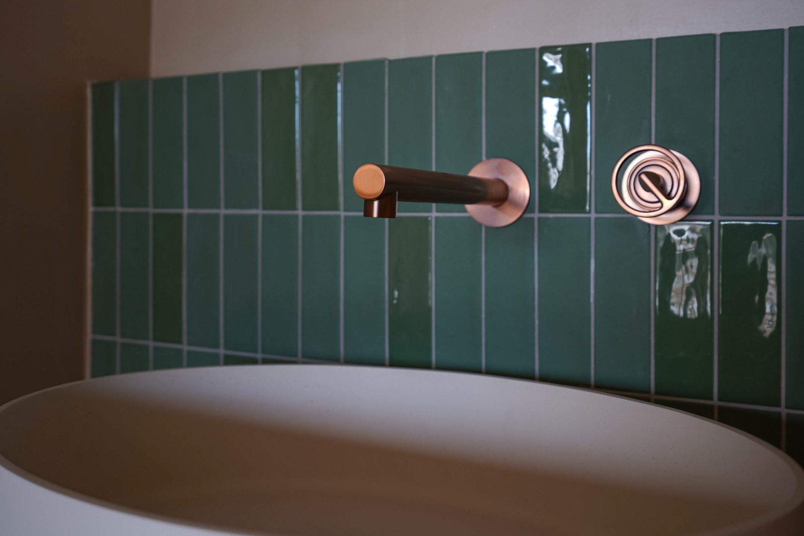 Detail van de badkamer met een koperen inbouwkraan, glanzende verticale tegels in smaragdgroen en een matwitte opzetwastafel.