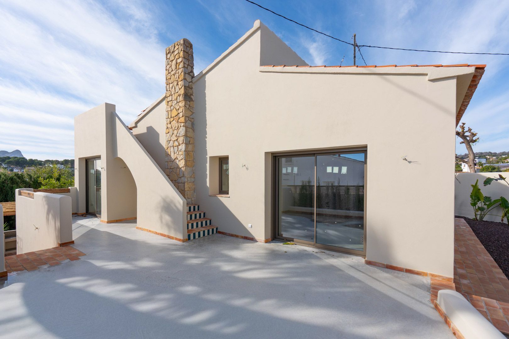 Exterieur van de villa met gepolijst betonnen terras, natuurstenen schoorsteen en trap met decoratieve tegels. Uitzicht op de Peñón de Ifach in de verte.