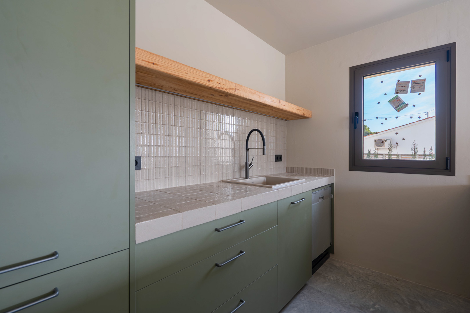 Gastenkeuken met saliegroene kasten, betegeld werkblad, massief houten plank en zwarte designkraan op een gepolijste betonvloer.