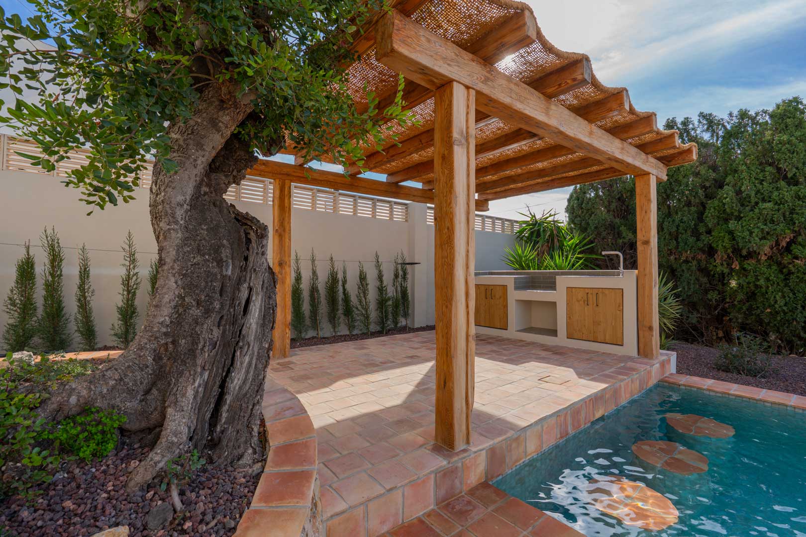 Houten pergola met een dak van natuurlijke vezels op terracotta tegels, voorzien van een buitenkeuken en een zwembad met onderwaterzitjes naast een oude olijfboom.