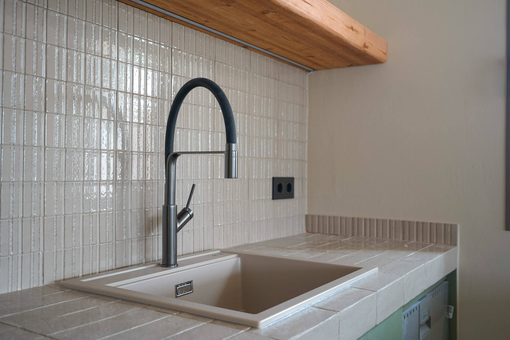 Keukendetail met een werkblad van porseleinen tegels en geïntegreerde spoelbak. Moderne flexibele kraan tegen een verticale mozaïek achterwand en massief houten plank.