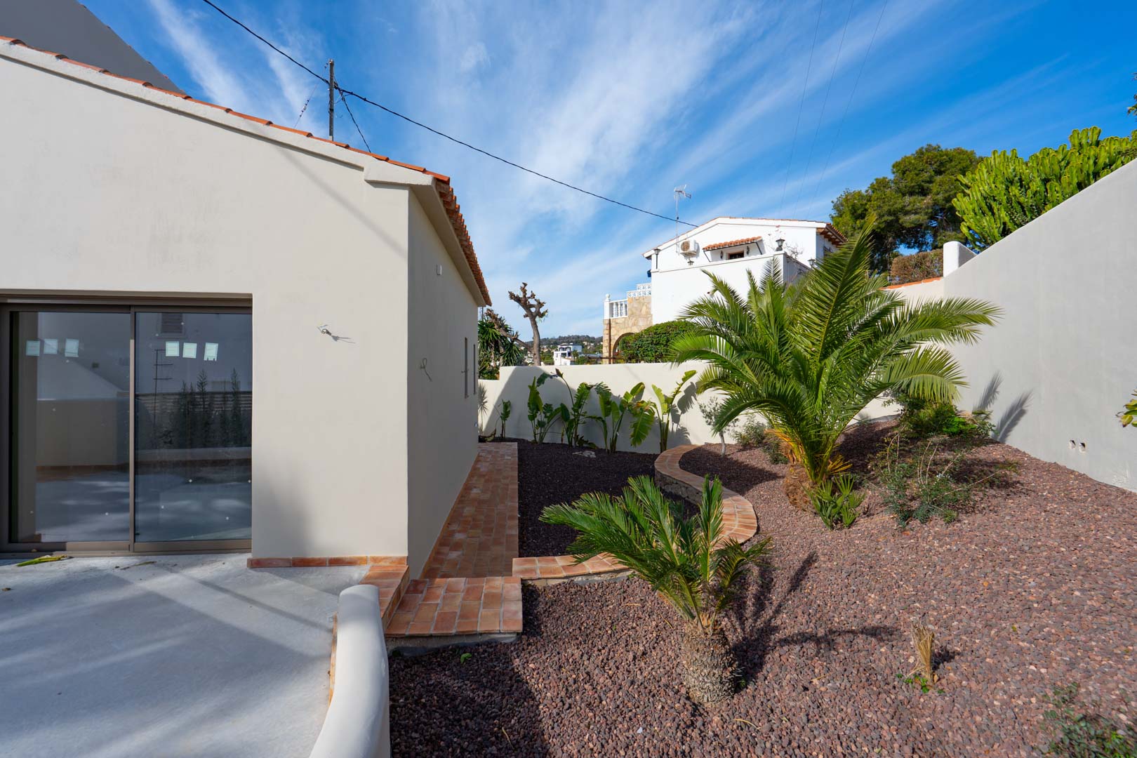 Mediterrane tuin met palmbomen en vulkanisch grind, omzoomd door terracotta paden en witte buitenmuren.