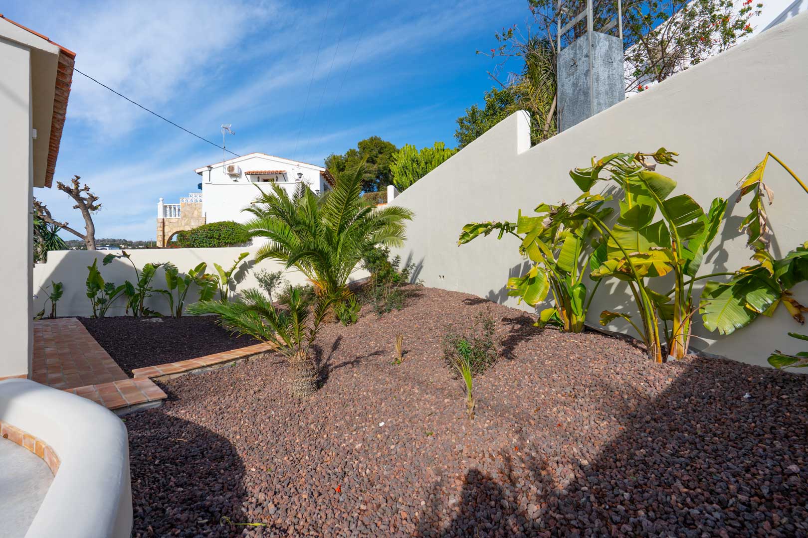 Mediterrane tuin met vulkanisch grind, palmen en bananenplanten naast een witte buitenmuur en een pad van terracotta tegels.
