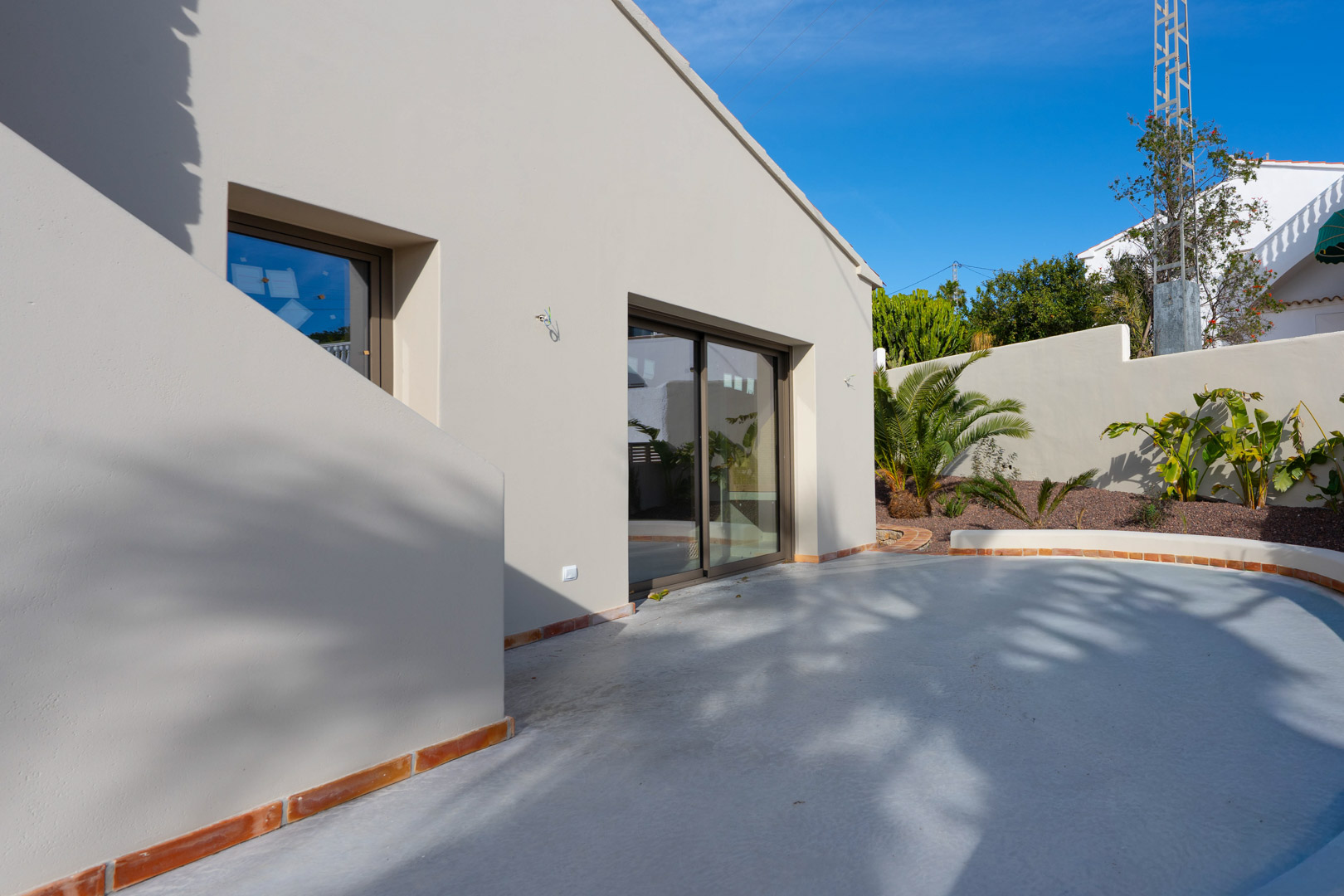 Minimalistische gevel in aardetinten met grote glazen schuifdeuren en een gebogen terras met terracotta randen en mediterrane beplanting.