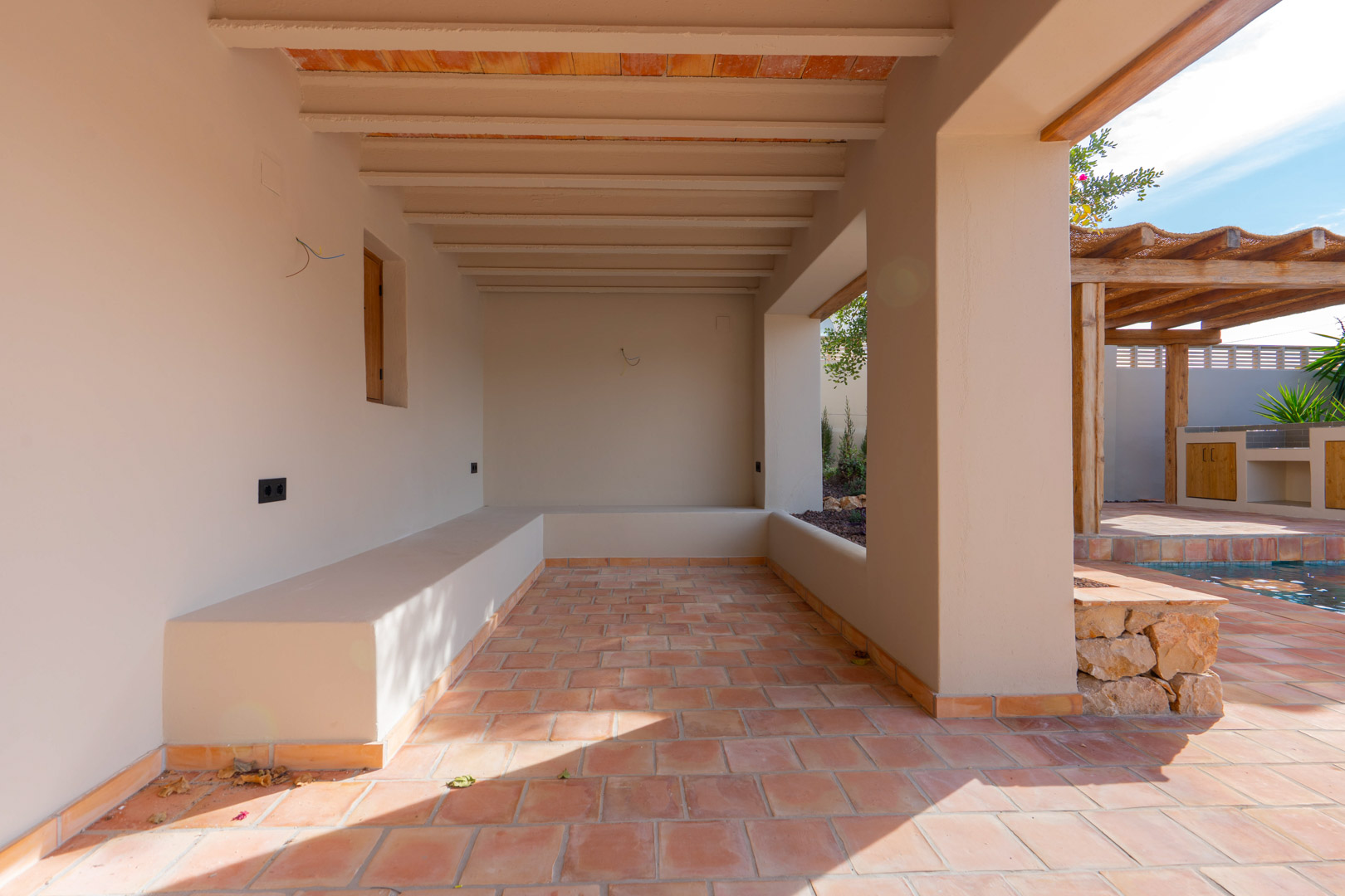 Overdekt terras met terracotta vloertegels, balkenplafond en ingebouwde zitbanken, grenzend aan het zwembad en de buitenkeuken.