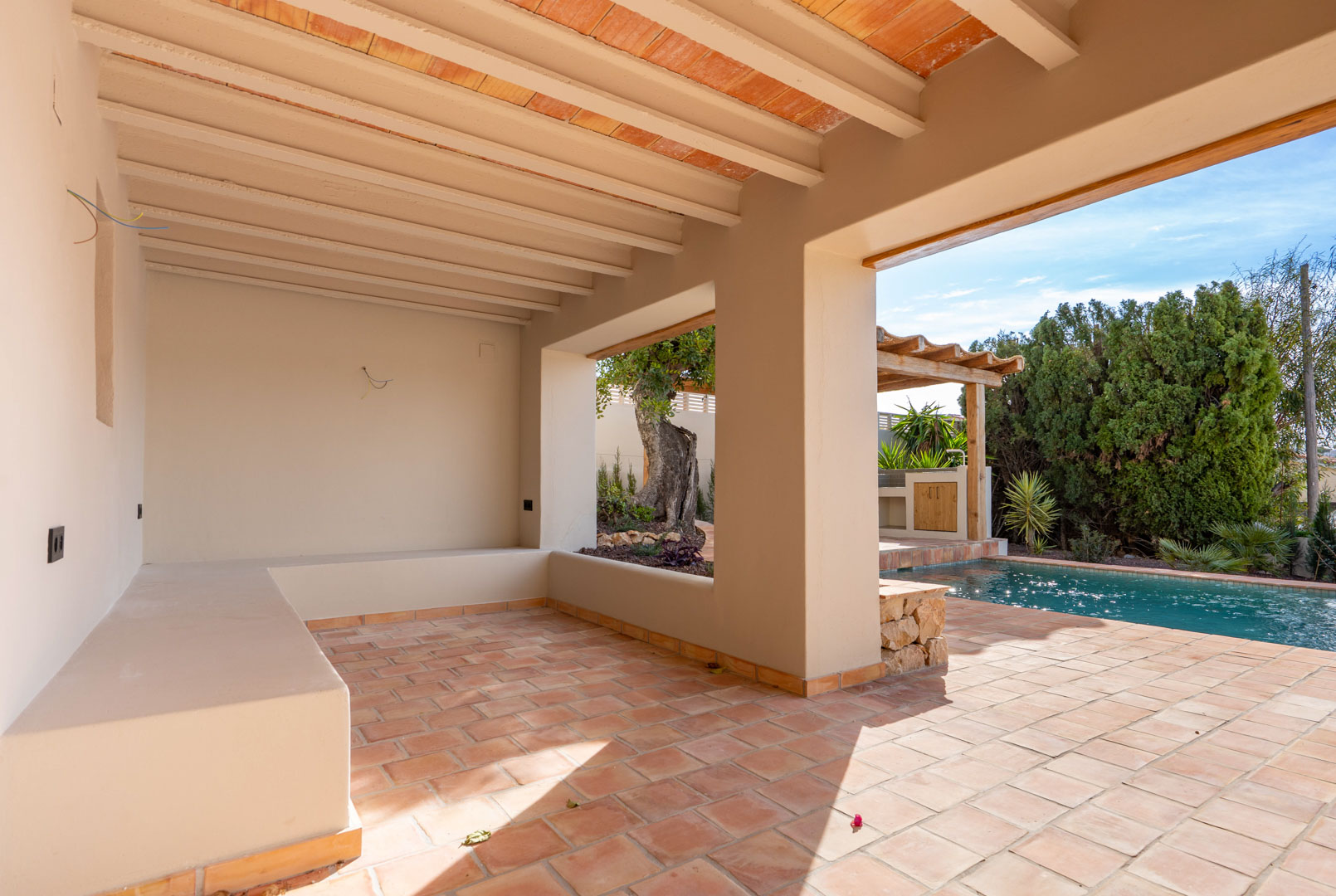 Overdekt terras met terracotta vloertegels, zichtbare plafondbalken en een gemetselde bank, met uitzicht op het zwembad en de pergola.