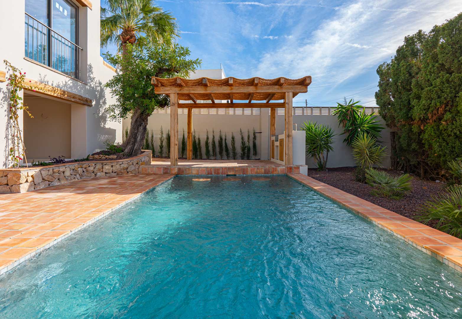 Rechthoekig zwembad met terracotta tegels, een houten pergola en een mediterrane tuin met natuurstenen muren, palmbomen en diverse groene beplanting.