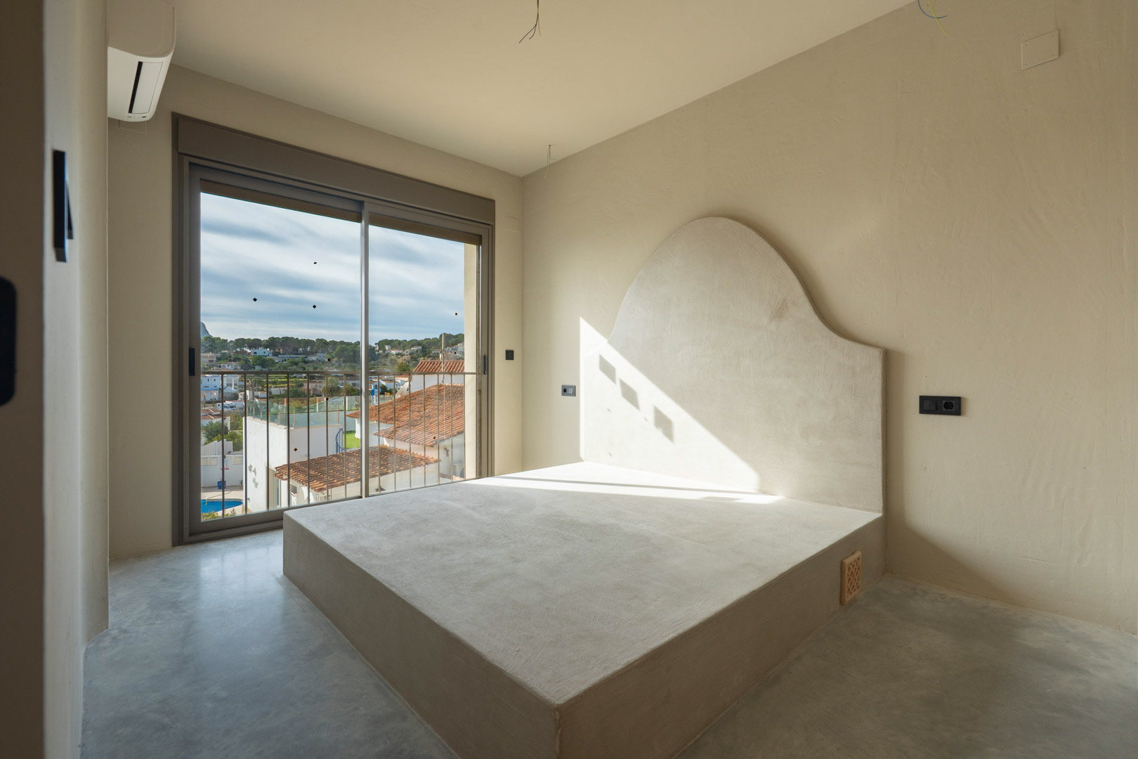 Slaapkamer met een ingebouwd bedframe en gebogen hoofdbord van klei, gepolijste betonvloer en schuifpui naar een balkon met uitzicht op de omgeving.