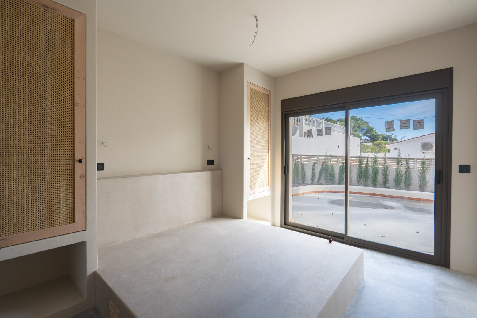 Slaapkamer met een ingebouwd bedplatform van gepolijst beton, maatwerkkasten met rotan panelen en een schuifpui die toegang biedt tot het terras.