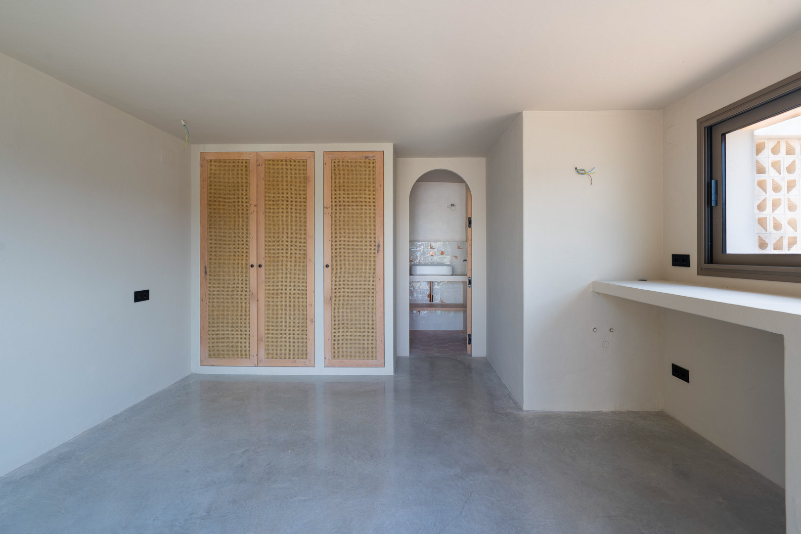 Slaapkamer met eikenhouten inbouwkasten met vlechtwerk, gepolijste betonvloer en een ronde toog die toegang biedt tot de aangrenzende badkamer.