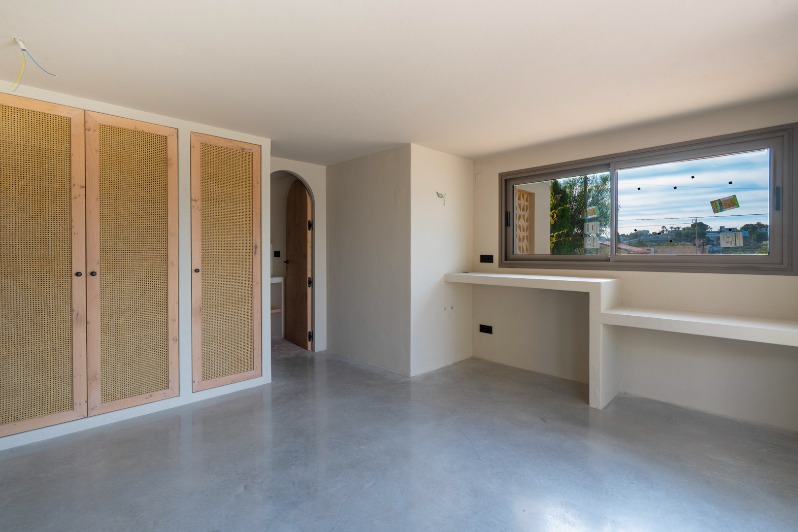 Slaapkamer met ingebouwde kasten met vlechtwerk, gepolijste betonvloer en een vast bureau onder het raam naast een karakteristieke ronde toog.