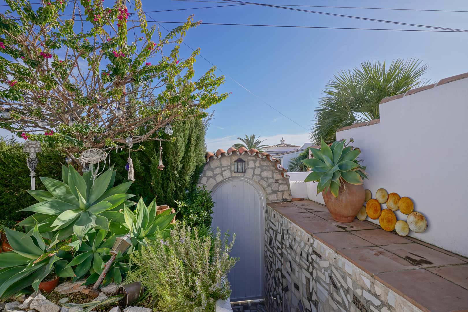 Buiteningang met een boog van natuursteen, lichtgrijze poort en mediterrane beplanting zoals agaves en bougainvillea langs witgestuukte muren.