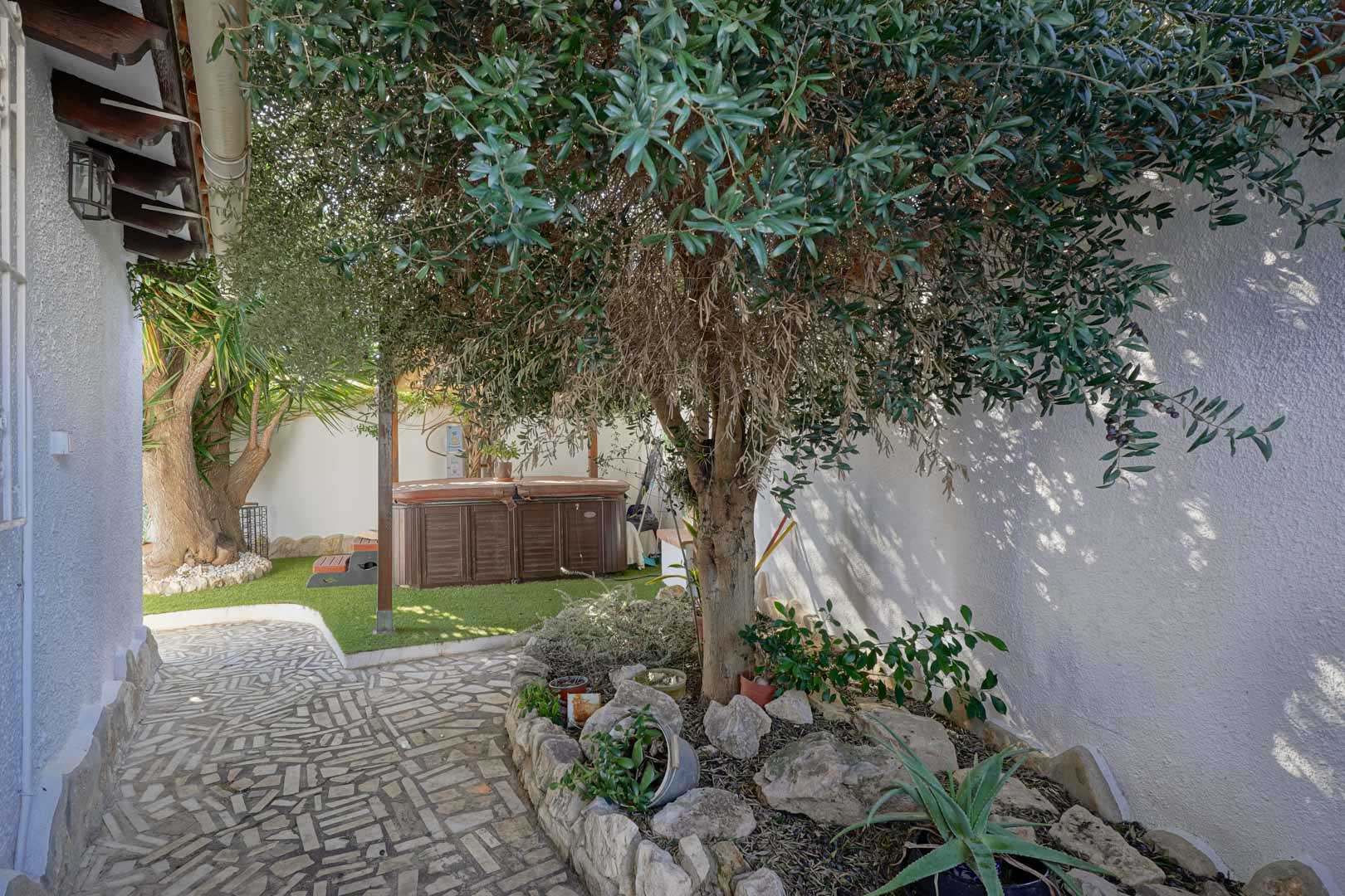 Buitenterras met privéjacuzzi op kunstgras, omgeven door mediterrane beplanting en een pad van natuursteen langs witte muren.