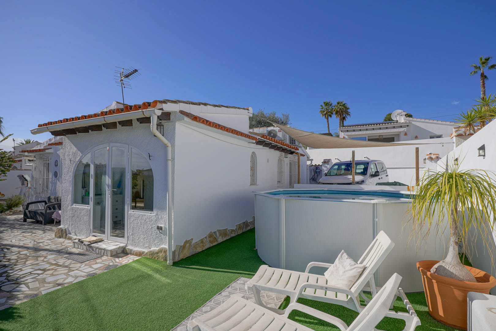 Exterieur van de witte villa met glazen veranda, een opzet zwembad op kunstgras en een zonneterras met witte ligbedden.