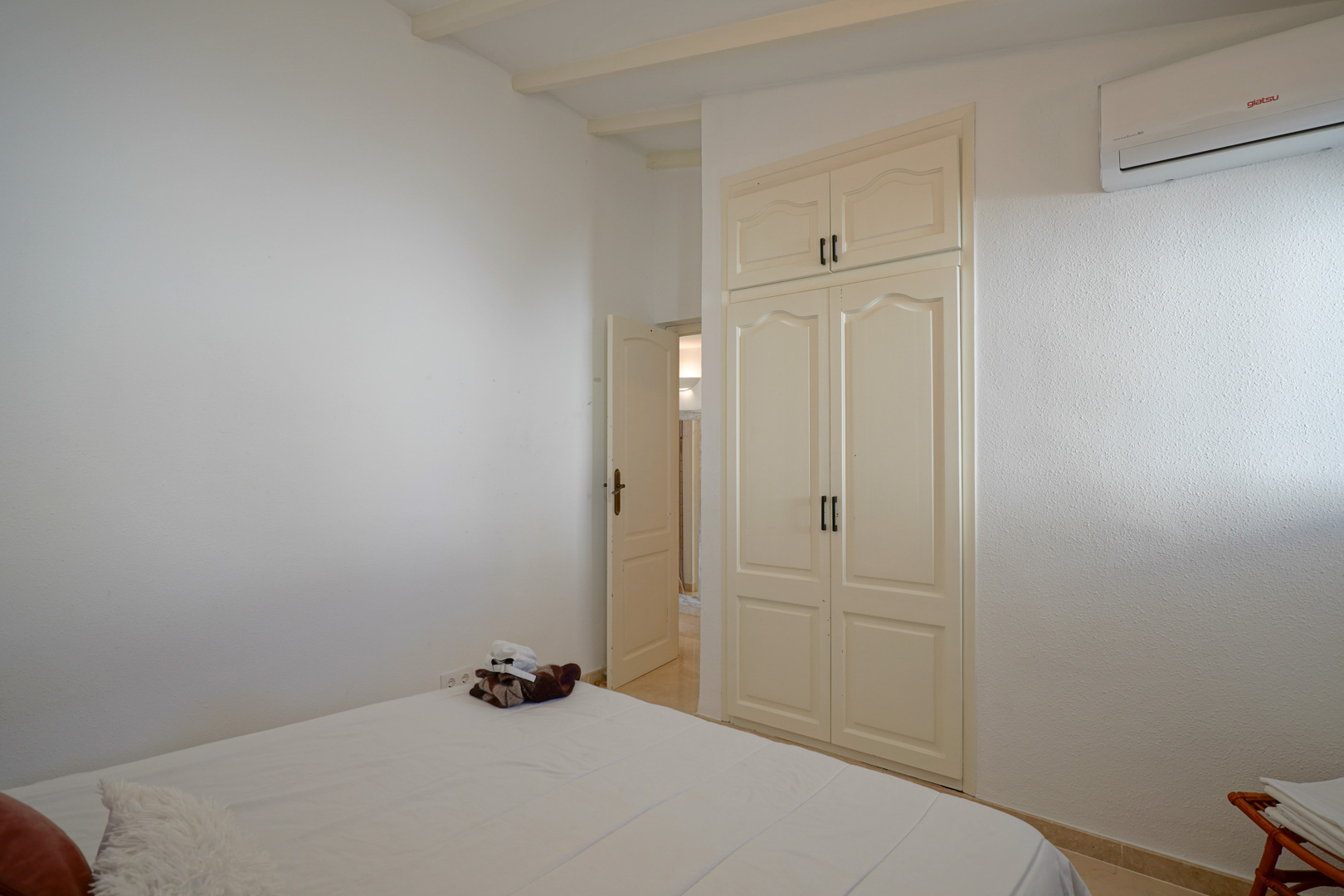 Lichte slaapkamer met een ingebouwde kledingkast met klassieke deuren, zichtbare plafondbalken en een wandgemonteerde airconditioning.