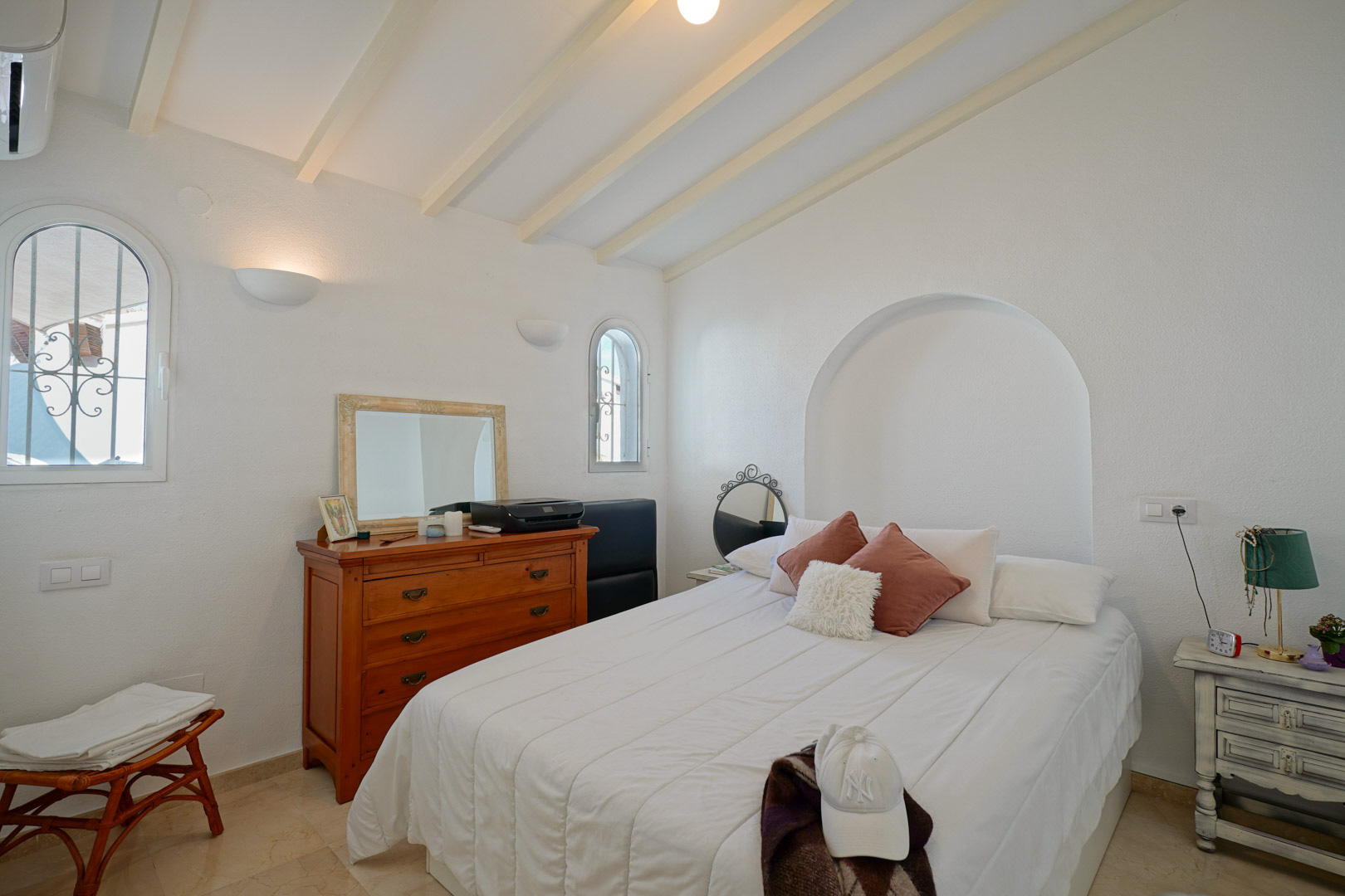 Lichte slaapkamer met witgeschilderde plafondbalken, boogramen en een decoratieve nis. Voorzien van airconditioning en een marmeren vloer.