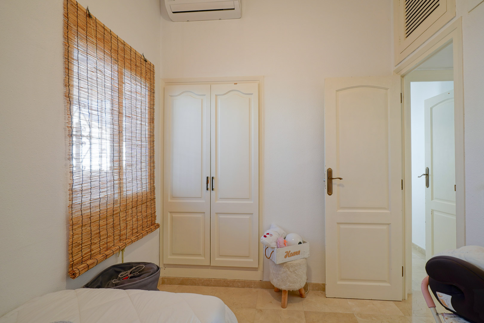 Lichte slaapkamer voorzien van een witte inbouwkast, split-airconditioning en raam met bamboe rolgordijn op een tegelvloer.