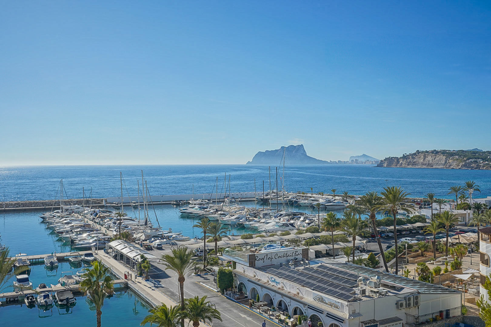 Panoramisch uitzicht op de jachthaven van Moraira met de Peñón de Ifach aan de horizon, wat de mediterrane kustsfeer en de afgemeerde jachten benadrukt.