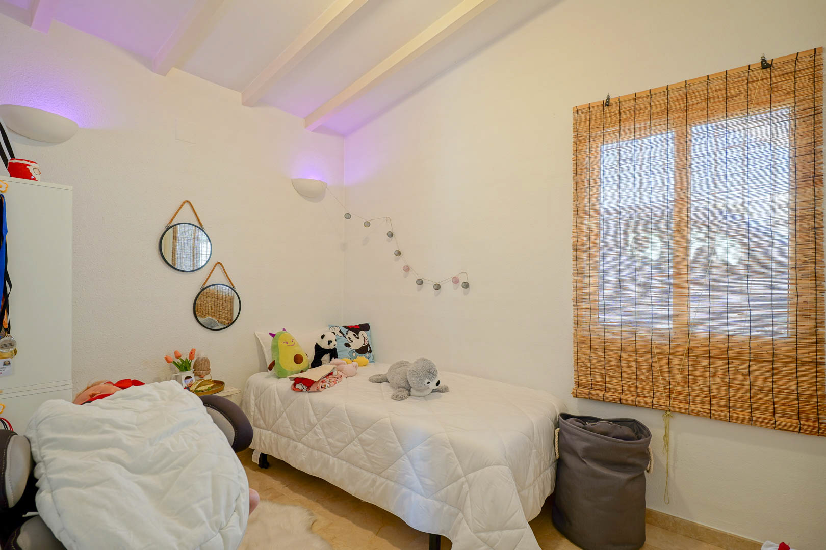 Sfeervolle slaapkamer met wit geschilderd balkenplafond, eenpersoonsbed, decoratieve spiegels en een rolgordijn van natuurlijke vezels.