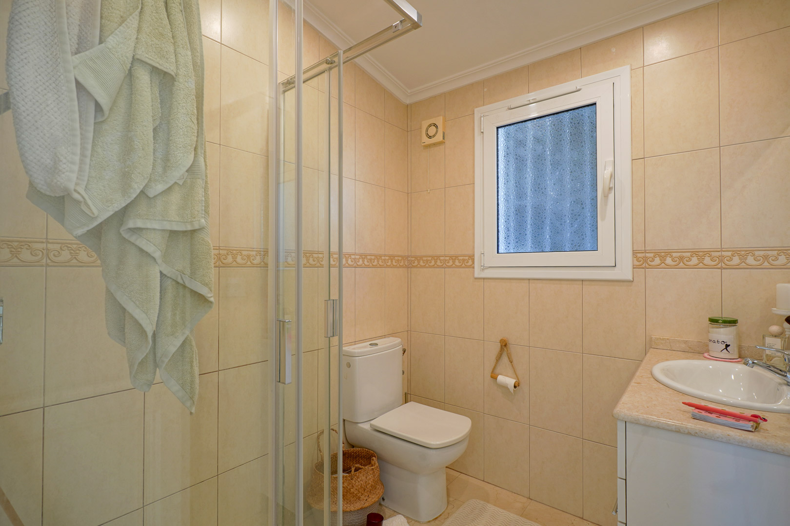Volledige badkamer met crème betegelde wanden, een glazen douchecabine, wit sanitair en een wastafelmeubel met een marmerlook blad.