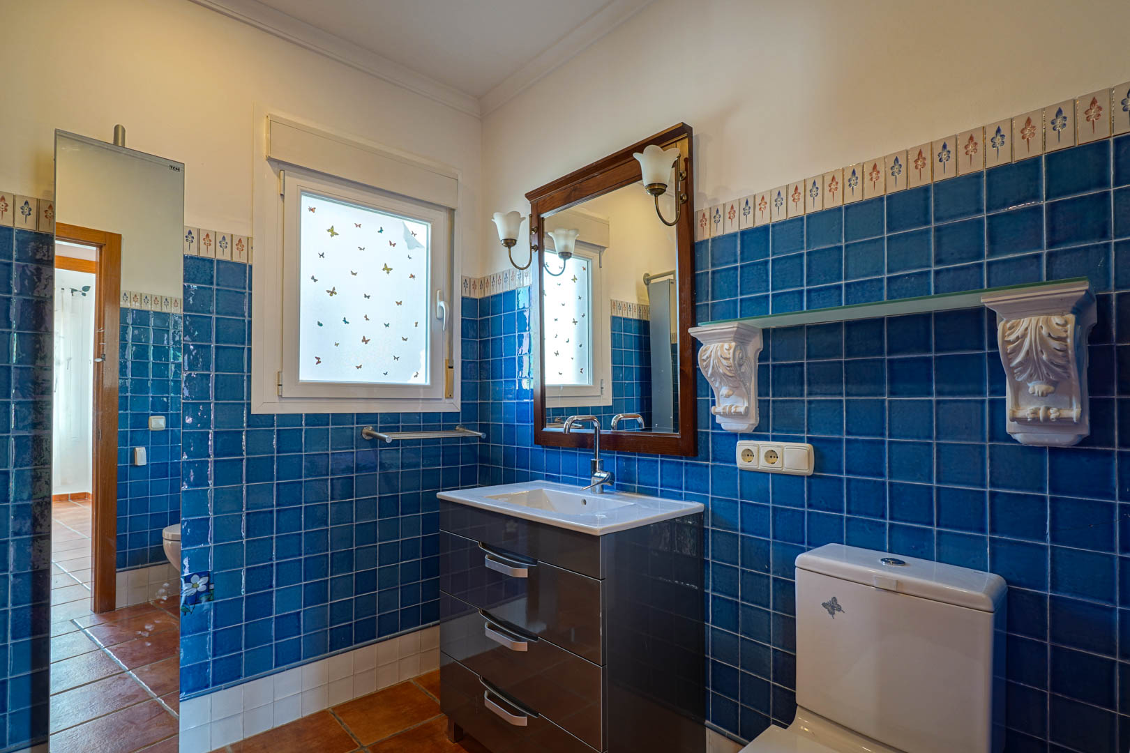 Badkamer met blauwe wandtegels en decoratieve rand, modern wastafelmeubel, spiegel met houten lijst en authentieke terracotta vloertegels.