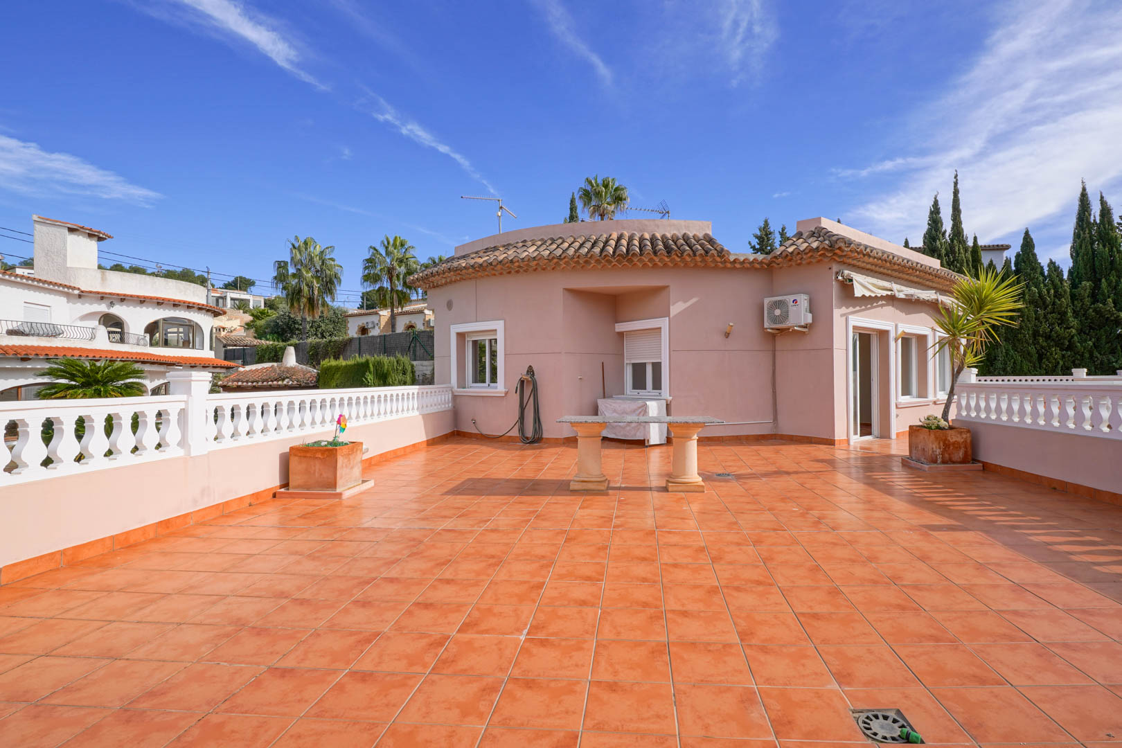 Ruim dakterras met terracotta tegels, een witte balustrade en een stenen tafel, omgeven door mediterrane architectuur en een heldere hemel.