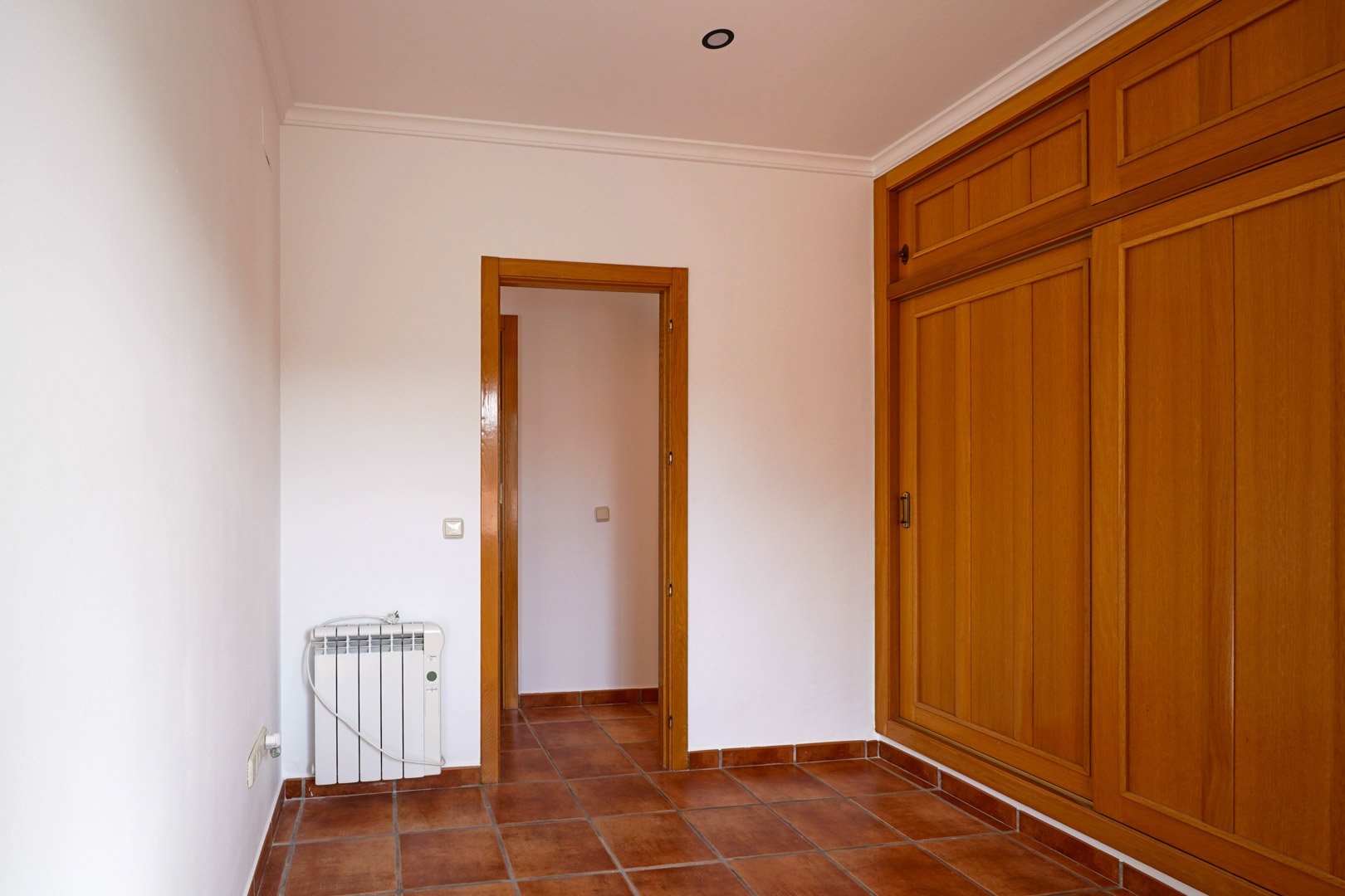 Slaapkamer met een grote ingebouwde houten kledingkast, rustieke terracotta vloertegels en een elektrische radiator. De ruimte heeft witte muren en sierlijsten aan het plafond.
