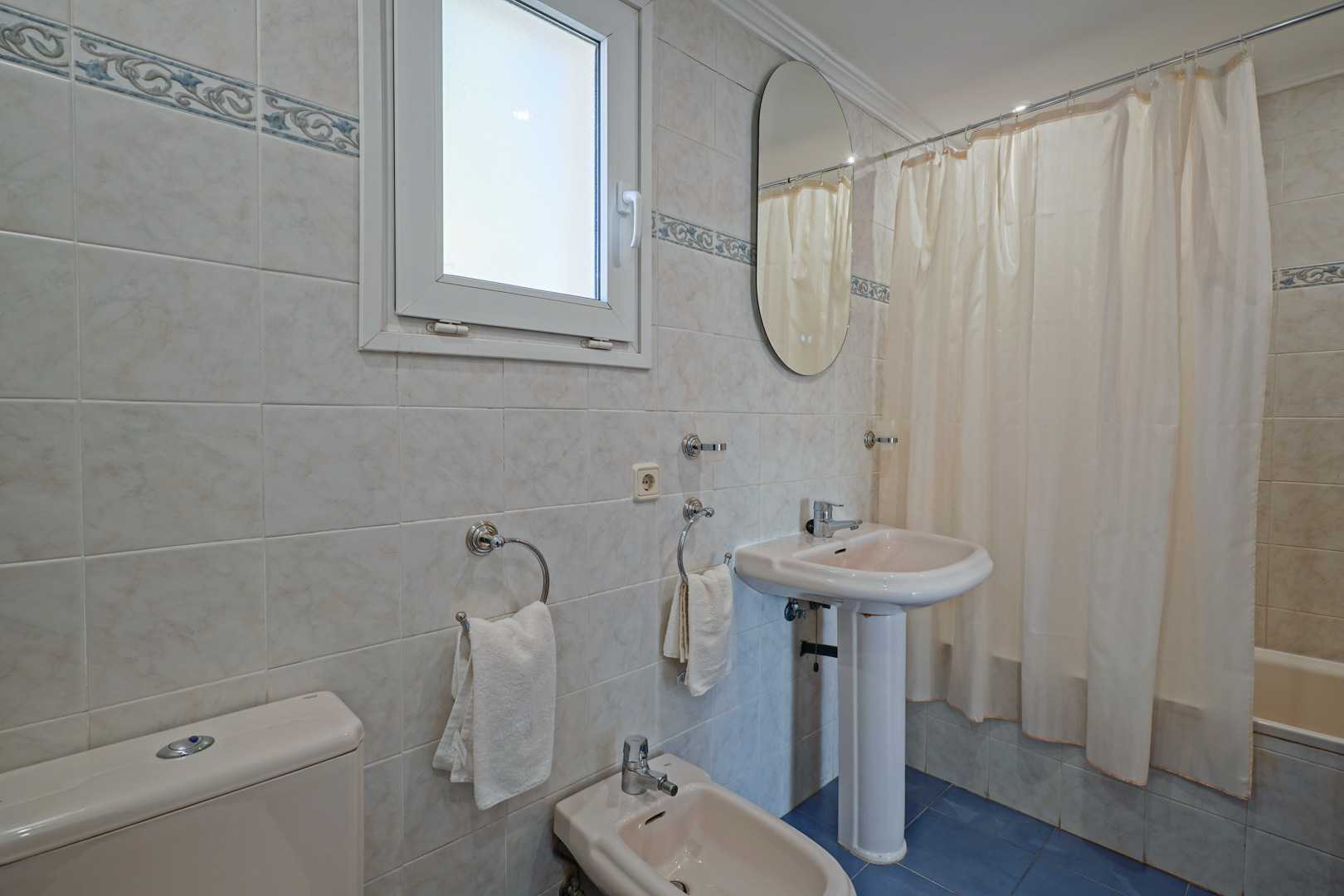 Familiale badkamer met crèmekleurig sanitair, waaronder een bidet en kolomwastafel. Voorzien van een ligbad, blauwe vloertegels en een raam voor natuurlijke lichtinval.