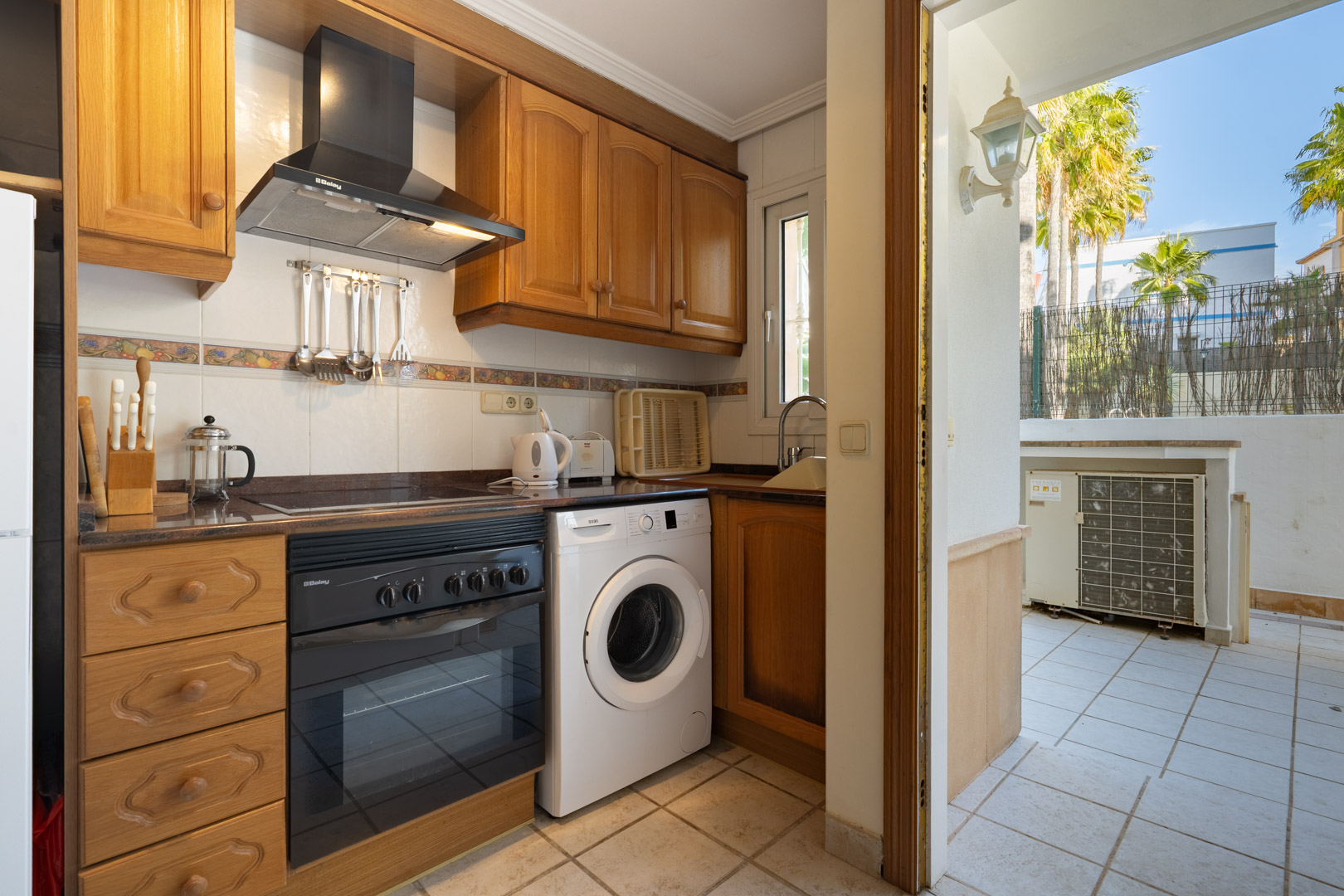 Functionele keuken met houten kasten, inbouwapparatuur en directe toegang tot een betegeld terras met uitzicht op palmbomen.