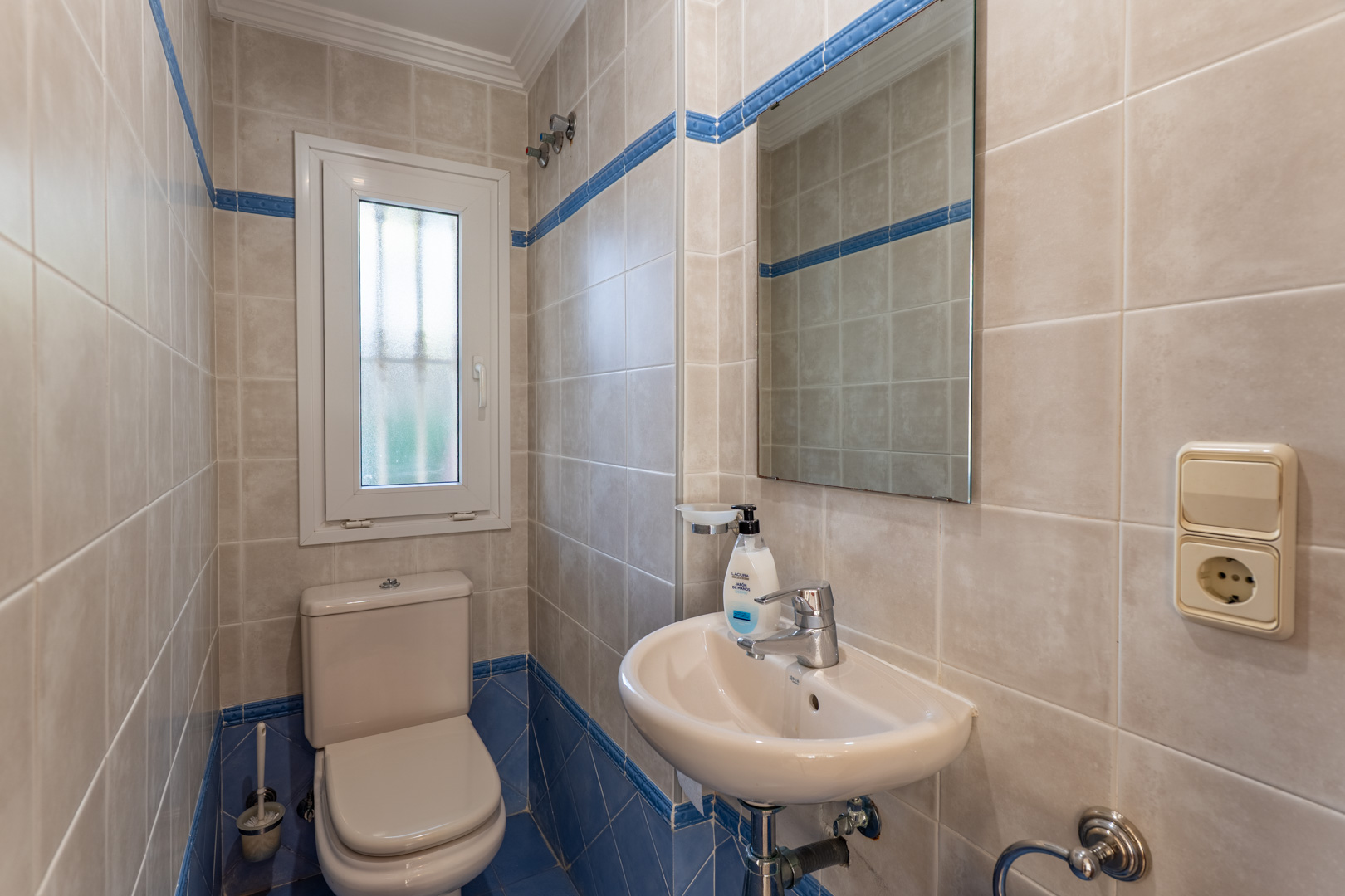 Gastentoilet met beige en blauwe wandtegels, voorzien van een wit porseleinen wastafel, spiegel en een raam voor natuurlijke ventilatie.