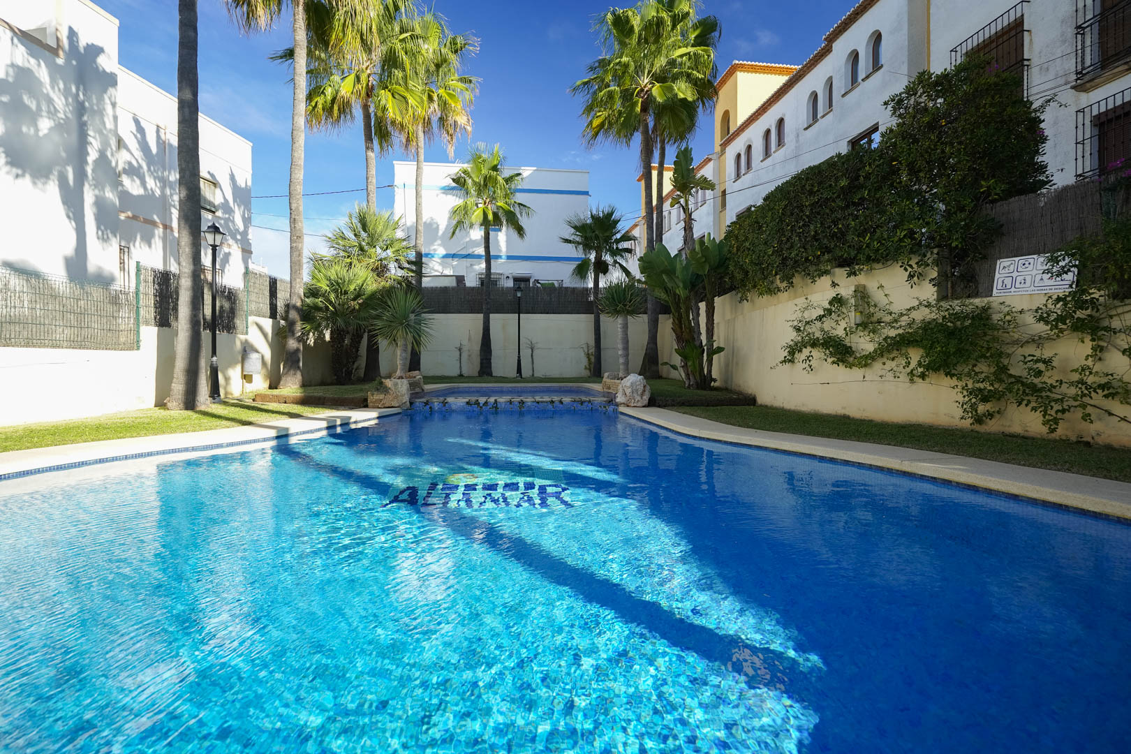 Gemeenschappelijk zwembad met blauwe tegels, omgeven door palmbomen en gazon. De buitenruimte biedt een rustige en zonnige omgeving in een exclusief complex in Jávea.