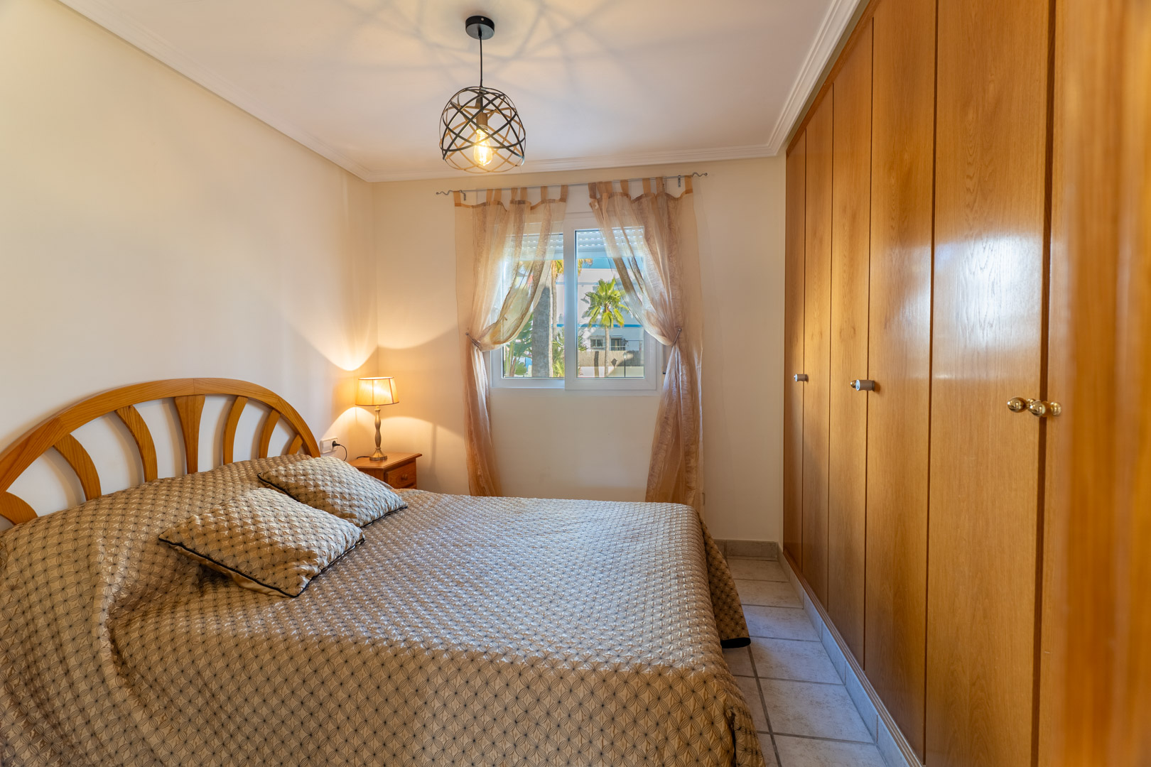 Lichte slaapkamer met ruime houten inbouwkasten, een tweepersoonsbed en een raam met uitzicht op de palmbomen van de urbanisatie.