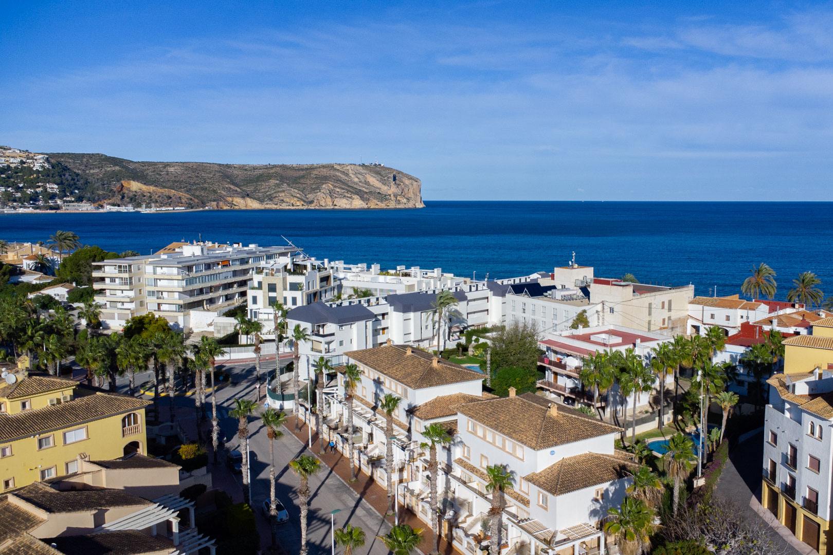 Panoramisch luchtfoto van Jávea met uitzicht op de Middellandse Zee en Cabo de San Antonio, gekenmerkt door traditionele pannendaken en straten omzoomd met palmbomen.