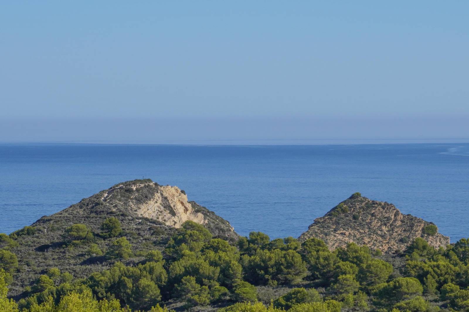 Panoramisch uitzicht op de Middellandse Zee, met rotsachtige kustheuvels en groene pijnboombossen onder een heldere horizon.