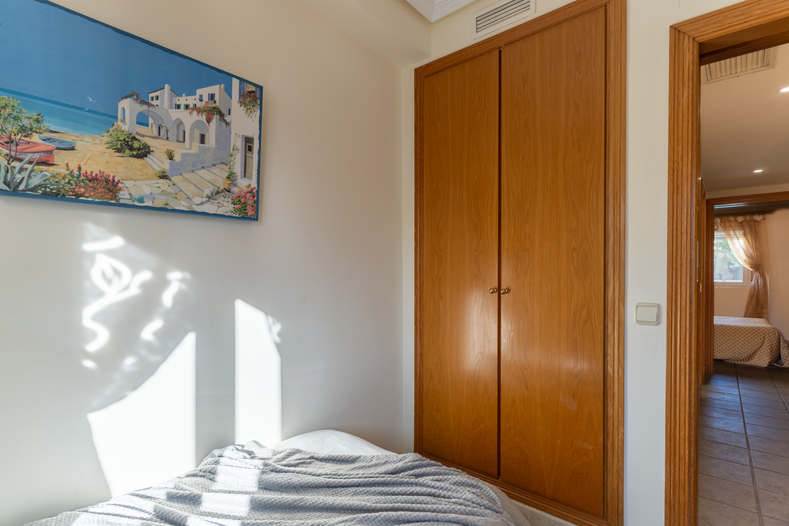 Slaapkamer met ingebouwde houten kledingkast en natuurlijk licht. De deuropening geeft toegang tot een betegelde gang en een aangrenzende kamer.