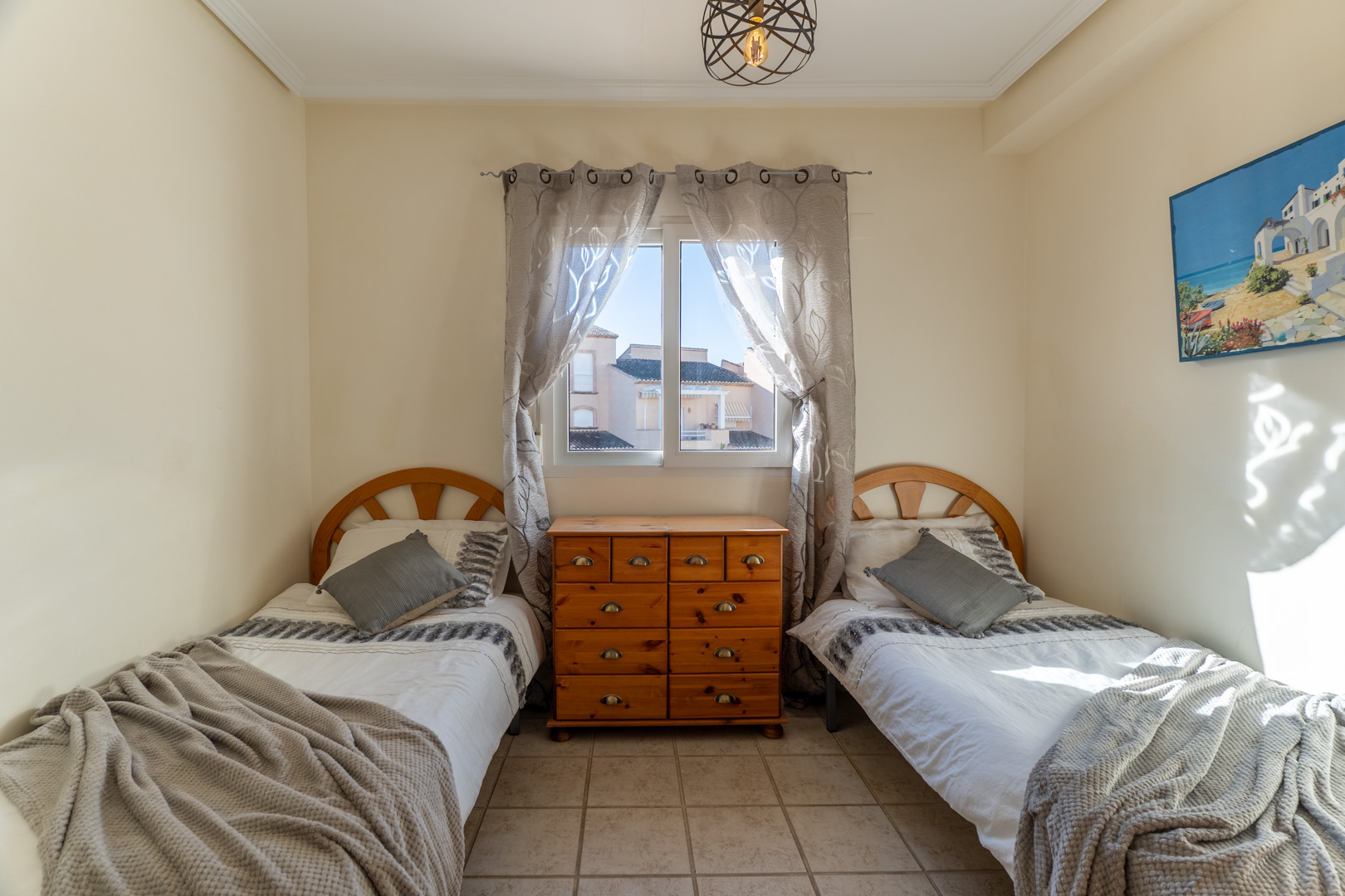 Slaapkamer met twee eenpersoonsbedden, houten hoofdeinden en een centrale ladekast. Voorzien van een tegelvloer, natuurlijk licht en een moderne hanglamp.