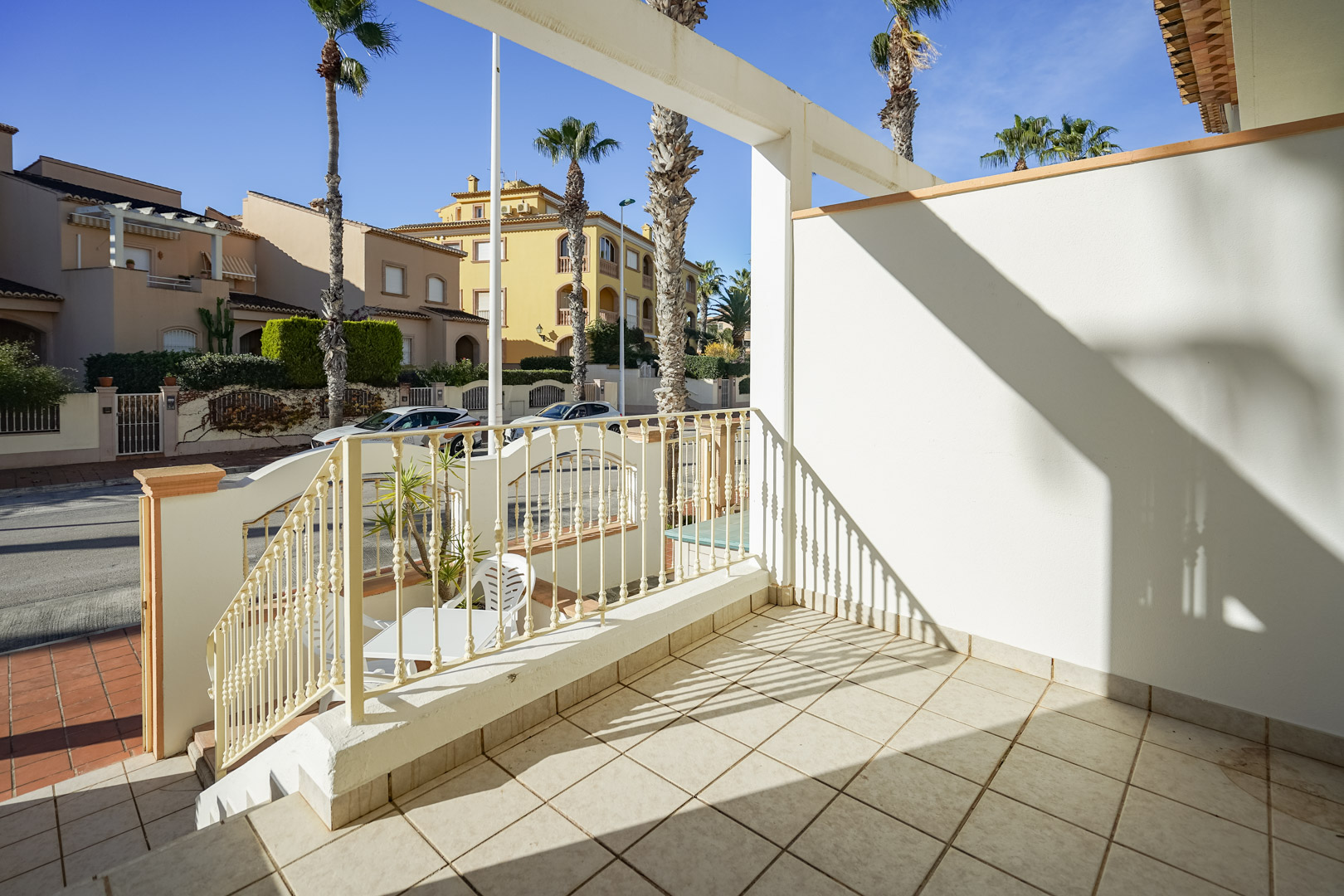 Verhoogd voorterras met keramische tegels en metalen balustrade, uitkijkend op de met palmbomen omzoomde straat in de woonwijk van Jávea.