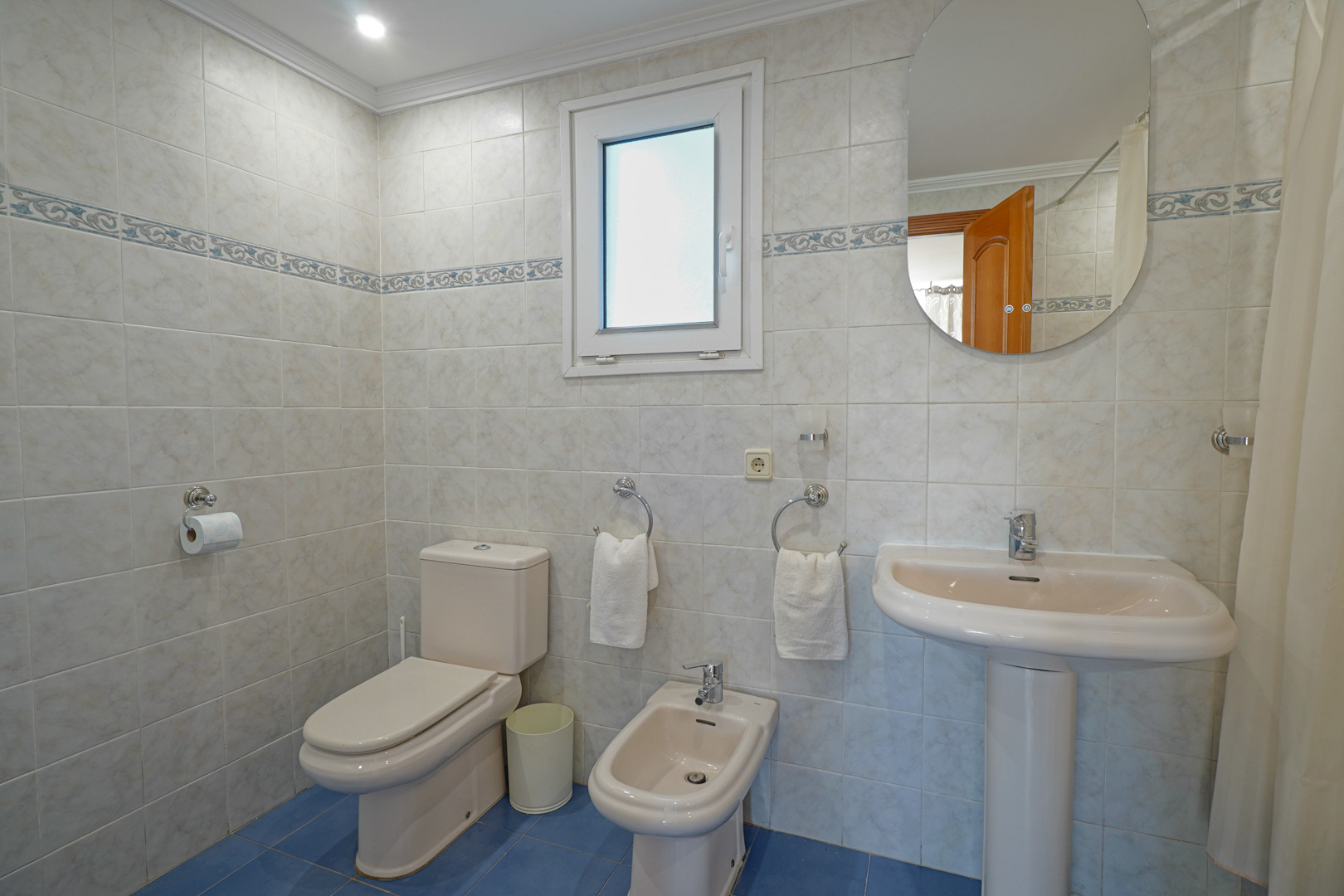 Volledige badkamer met lichte wandtegels, decoratieve bies en blauwe vloer. Voorzien van toilet, bidet, wastafel op zuil en een raam voor daglicht.