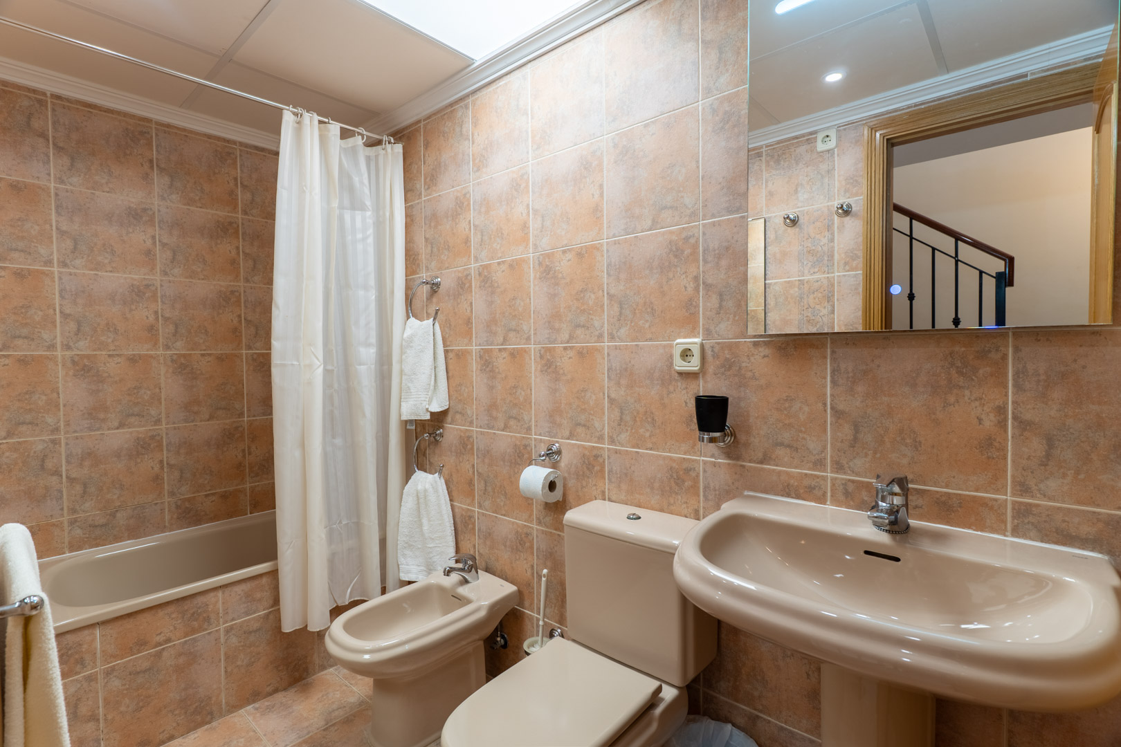 Volledige badkamer met ligbad, bidet, wastafel op zuil en aardetinten keramische tegels, verlicht door een dakraam.
