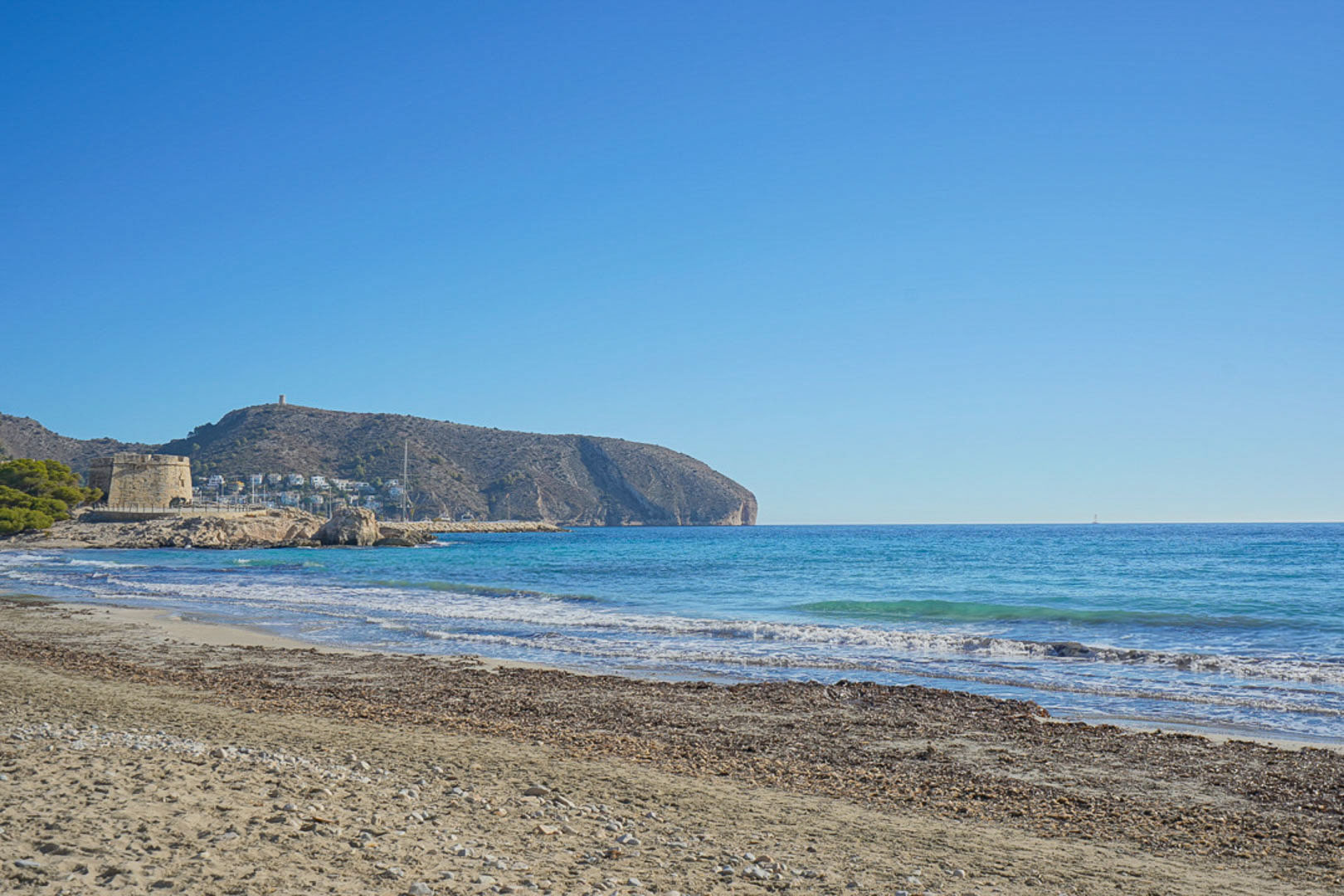 Uitzicht op het strand van Moraira met zijn historische toren en de Middellandse Zee onder een helderblauwe hemel.