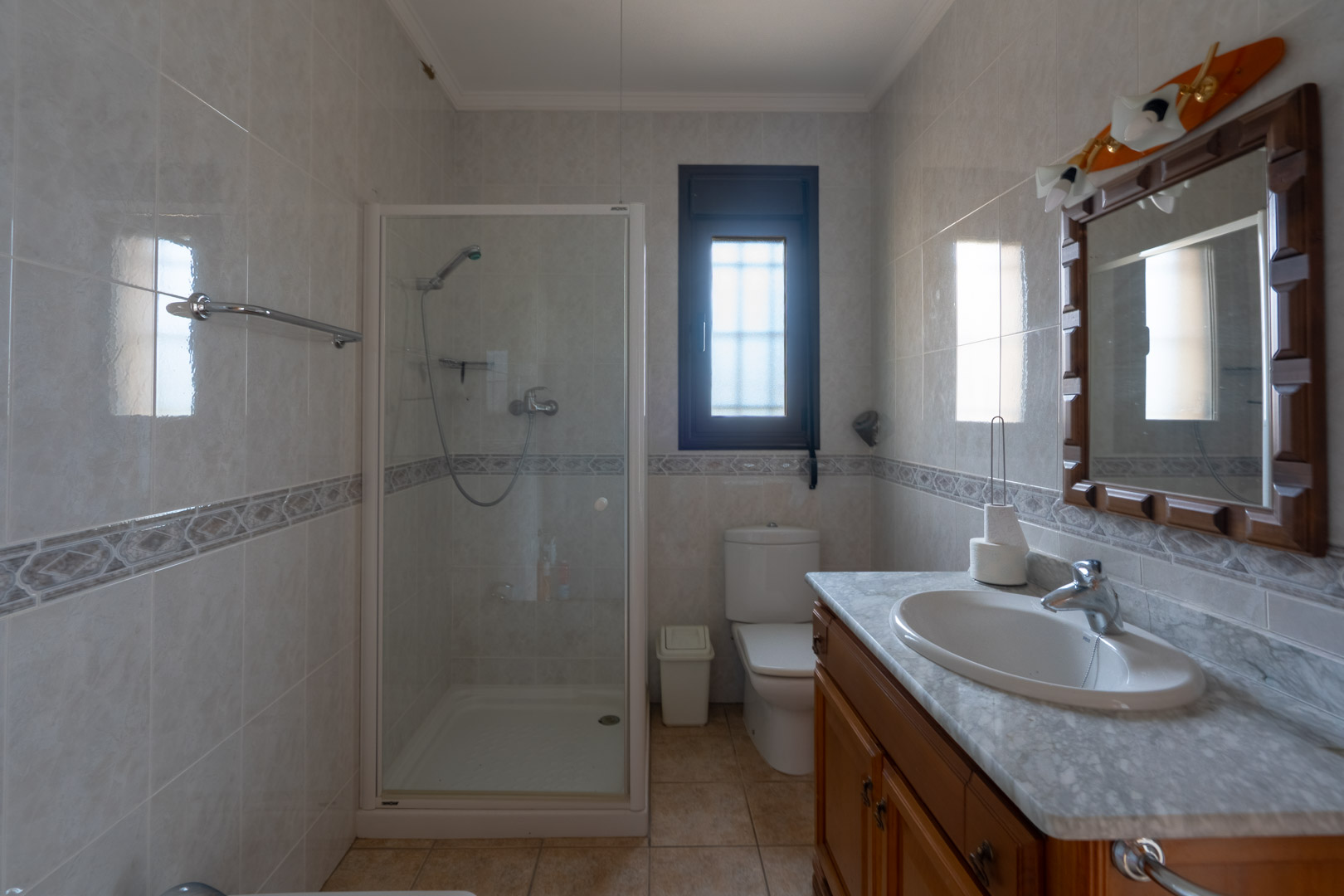 Badkamer met marmeren wastafelblad, houten meubel en glazen douchecabine. Betegelde wanden met een decoratieve bies en een raam met melkglas.