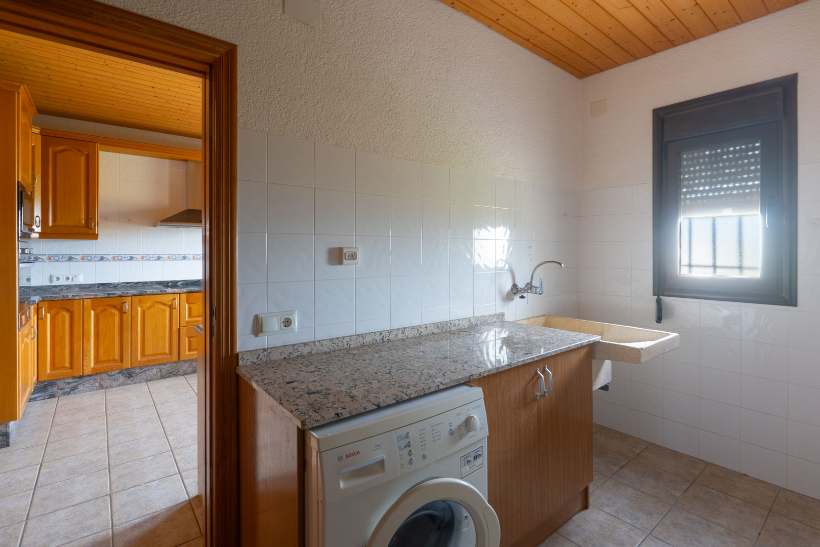 Bijkeuken met granieten werkblad, Bosch wasmachine en stenen spoelbak. Voorzien van een houten plafond en witte wandtegels, grenzend aan de keuken.